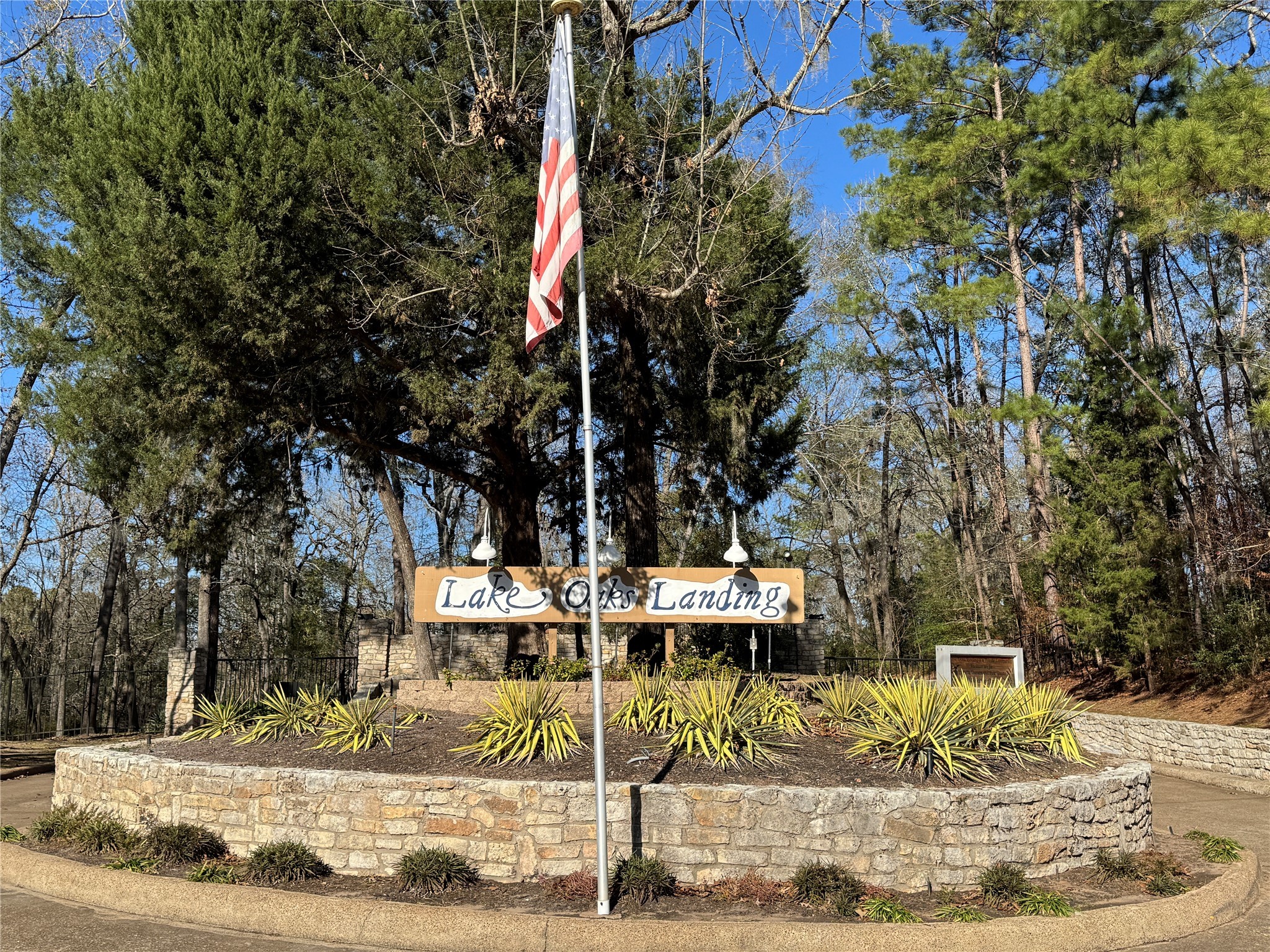 200 Harbor Run Drive Coldspring, TX 77331 - Photo 4 of 50 This photo shows the entrance to "Lake Oaks Landing," featuring a stone wall, landscaped greenery, and an American flag. The area is surrounded by lush trees, suggesting a serene and natural setting.