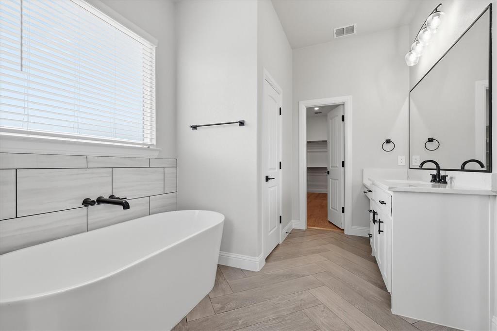 120 Grace Vw Road Weatherford, TX 76088 - Photo 17 of 40 Freestanding soaking tub with matte black wall-mounted faucet, herringbone wood-finish flooring, white vanity with stone countertop, and large rectangular mirror with modern lighting fixture