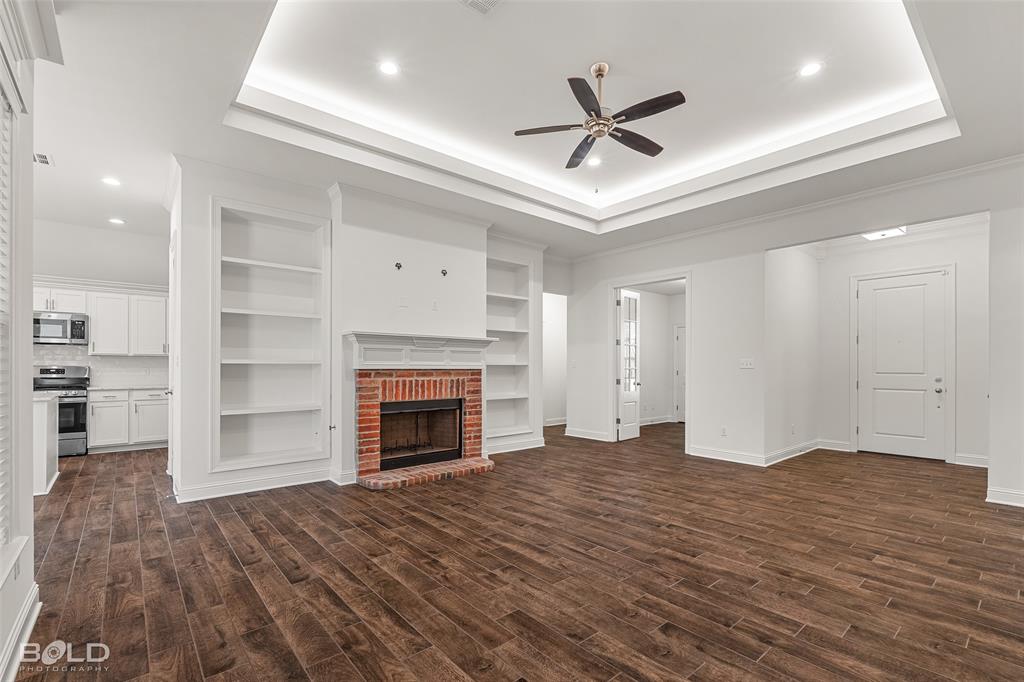 304 Prien Lk Loop Bossier City, LA 71111 - Photo 12 of 39 Spacious living area featuring wood-finish flooring, a brick fireplace with built-in shelving, a tray ceiling with recessed lighting and ceiling fan, and a pass-through to the kitchen with white cabinetry