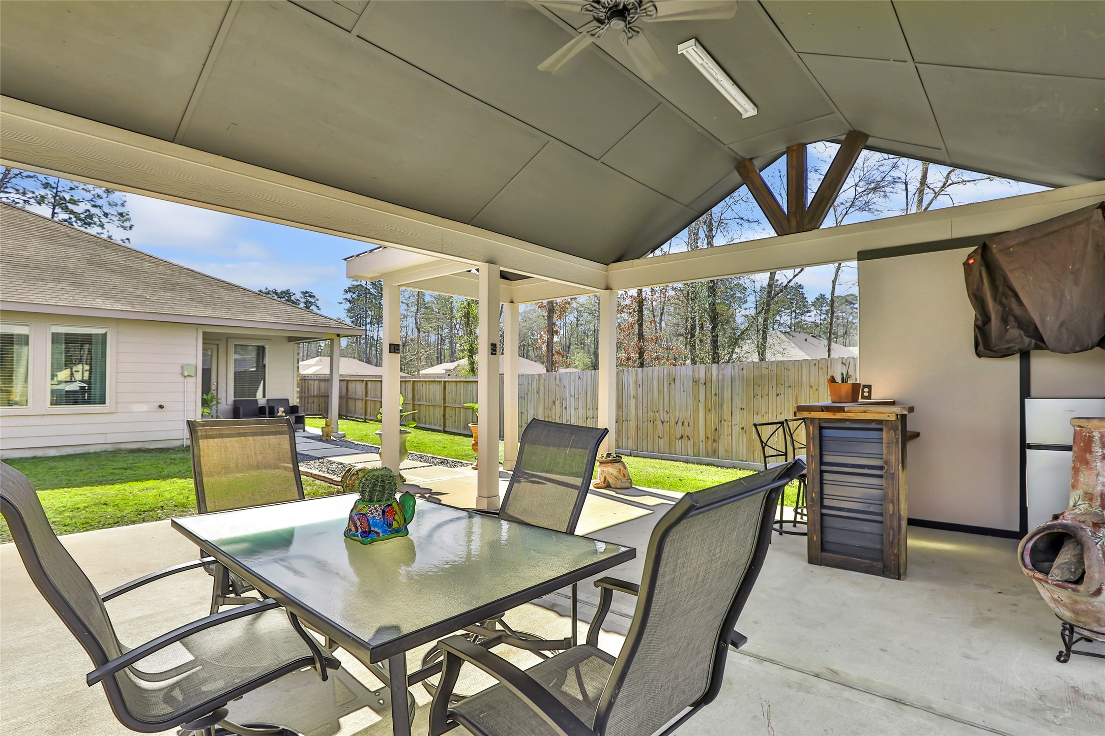811 Republic Road New Caney, TX 77357 - Photo 9 of 37 This photo showcases a covered patio with a ceiling fan, perfect for outdoor dining and relaxation. It includes a glass-top table, chairs, and a small bar area. The space opens to a fenced backyard with a well-maintained lawn, providing a private outdoor setting.