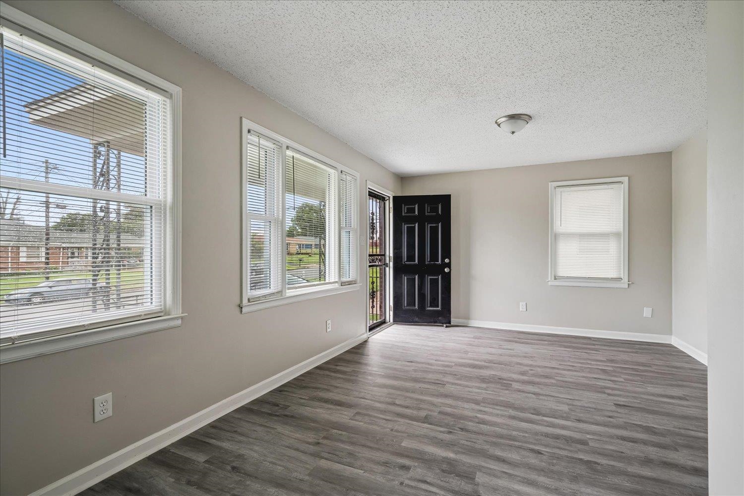 2518 Manchester Road Memphis, TN 38114 - Photo 7 of 23 Spacious interior with neutral wall paint, wood-finish flooring, multiple windows with blinds, a prominent black paneled entry door, and a ceiling-mounted light fixture