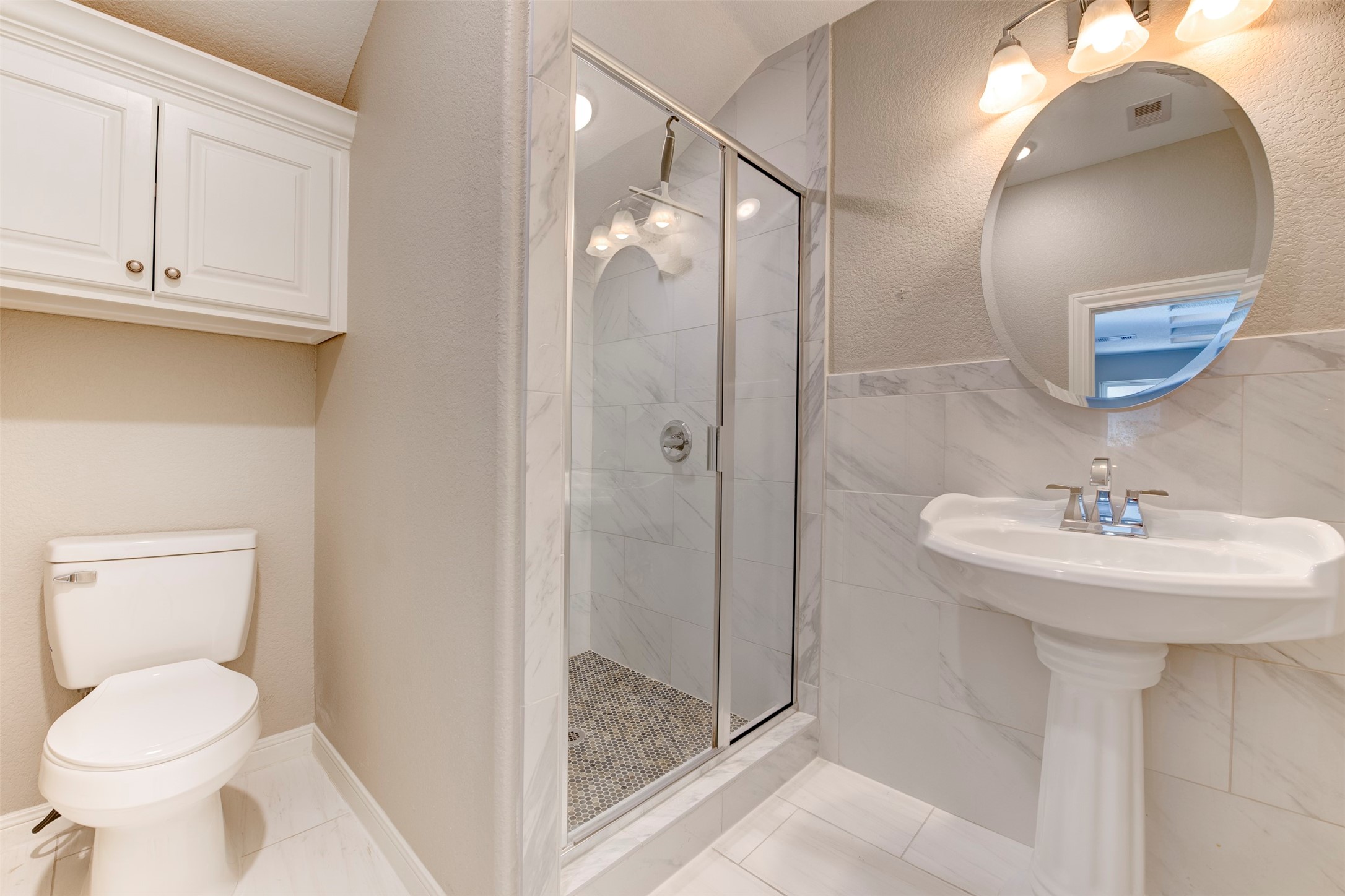 109 Dresden Street Houston, TX 77012 - Photo 28 of 43 This primary bathroom features a modern design with a glass-enclosed shower, pedestal sink, and a toilet. Elegant tile work and a round mirror add sophistication, while overhead lighting provides ample illumination.