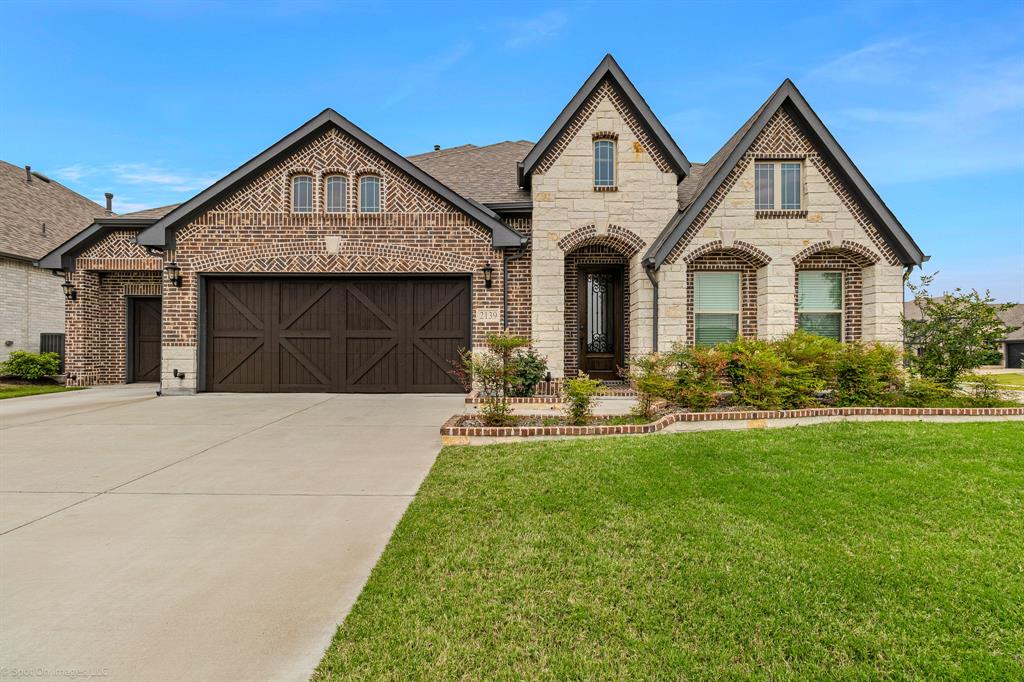 French country home with concrete driveway, 3 car garage, brick and stone exterior on a corner lot with rock and brick landscape border.