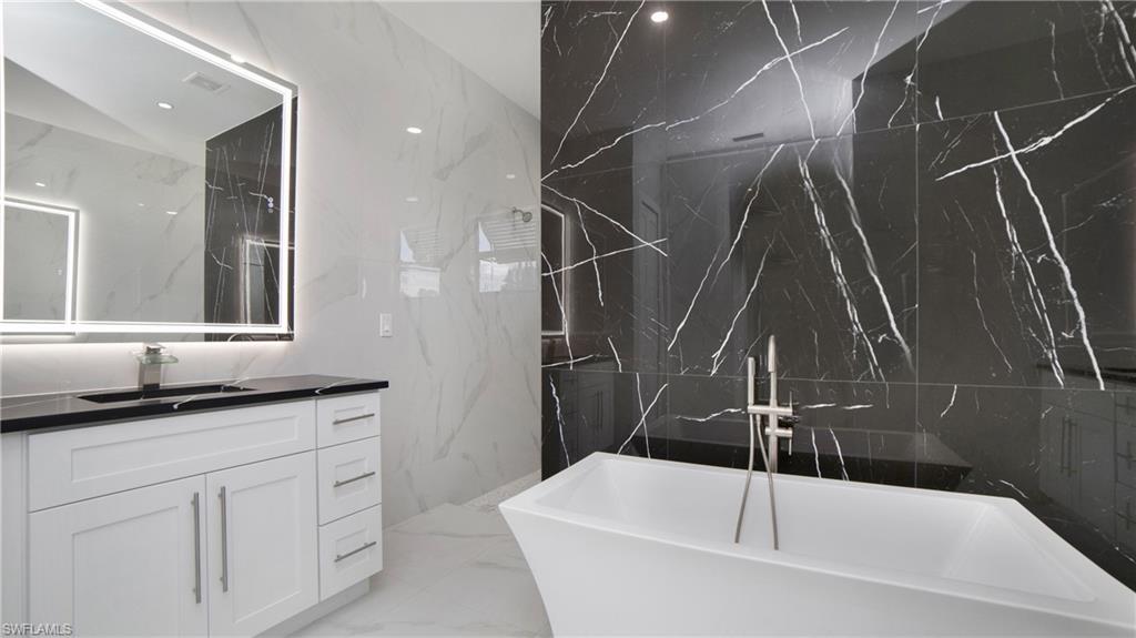 2343 Oil Well Road Naples, FL 34120 - Photo 19 of 46 Modern bathroom featuring a standalone soaking tub with floor-mounted faucet, black and white veined tile walls, a white vanity with shaker-style cabinetry, a dark countertop, and an illuminated mirror