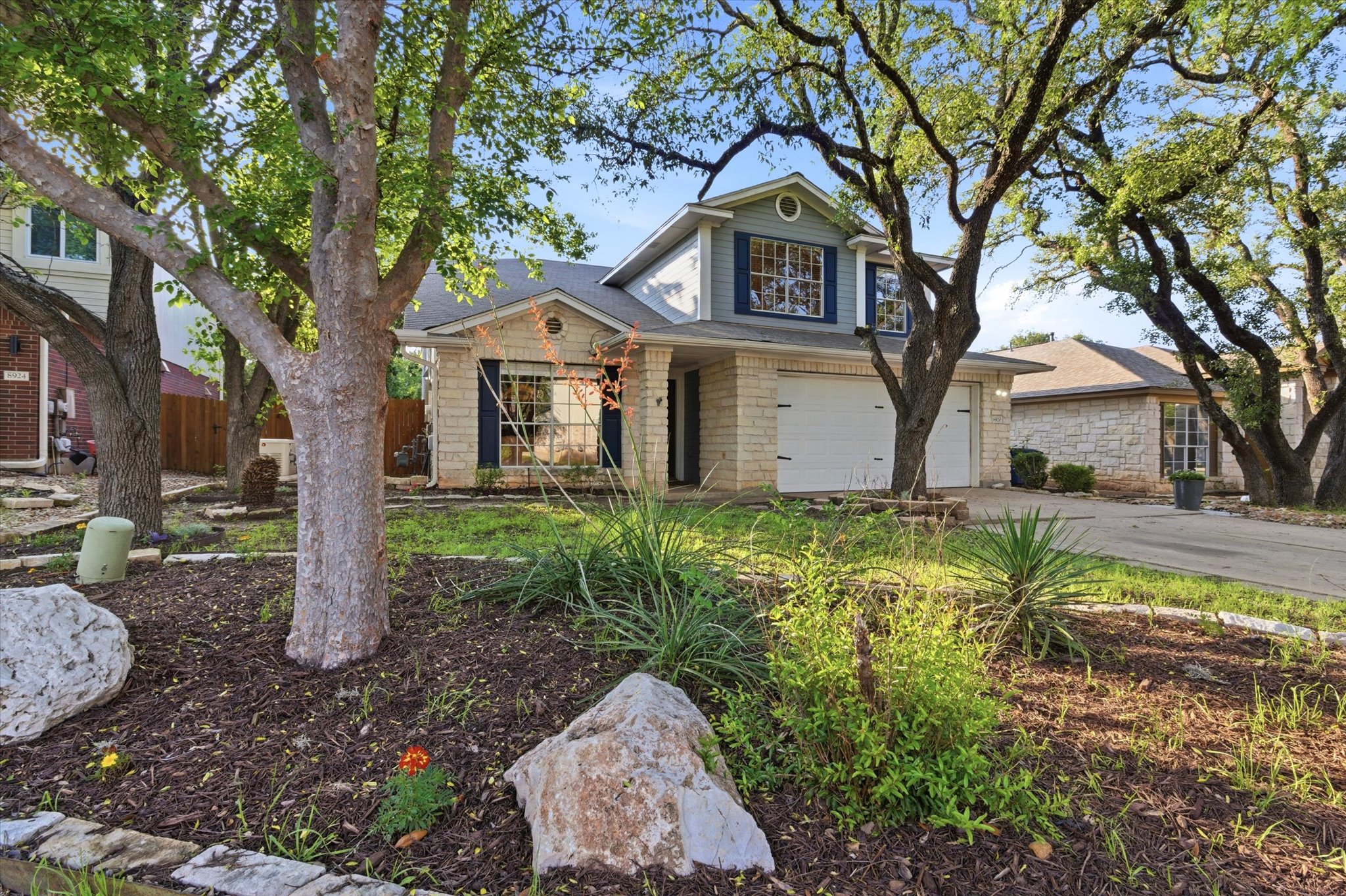 8920 West Hove Loop Austin, TX 78749 - Photo 34 of 39 Stone facade residence featuring a two-car garage, mature trees, and landscaping elements including decorative rocks and shrubs