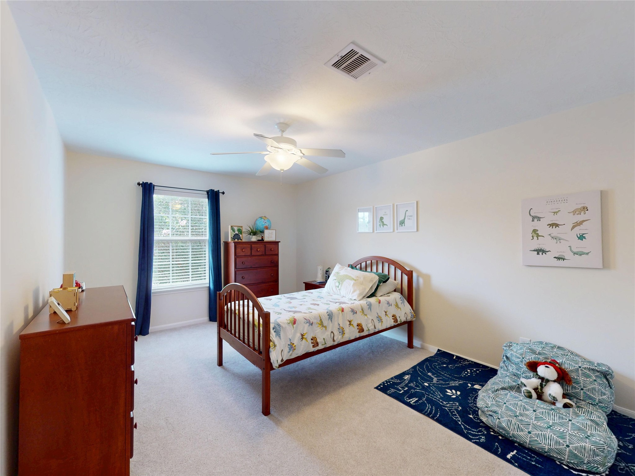 17402 Rainer Valley Lane Humble, TX 77346 - Photo 23 of 33 This bright, cozy bedroom features neutral walls and carpet, a ceiling fan, a single bed with dinosaur-themed bedding, and ample natural light from a window with dark curtains. Ideal for a child's room, it includes a dresser and playful decor.