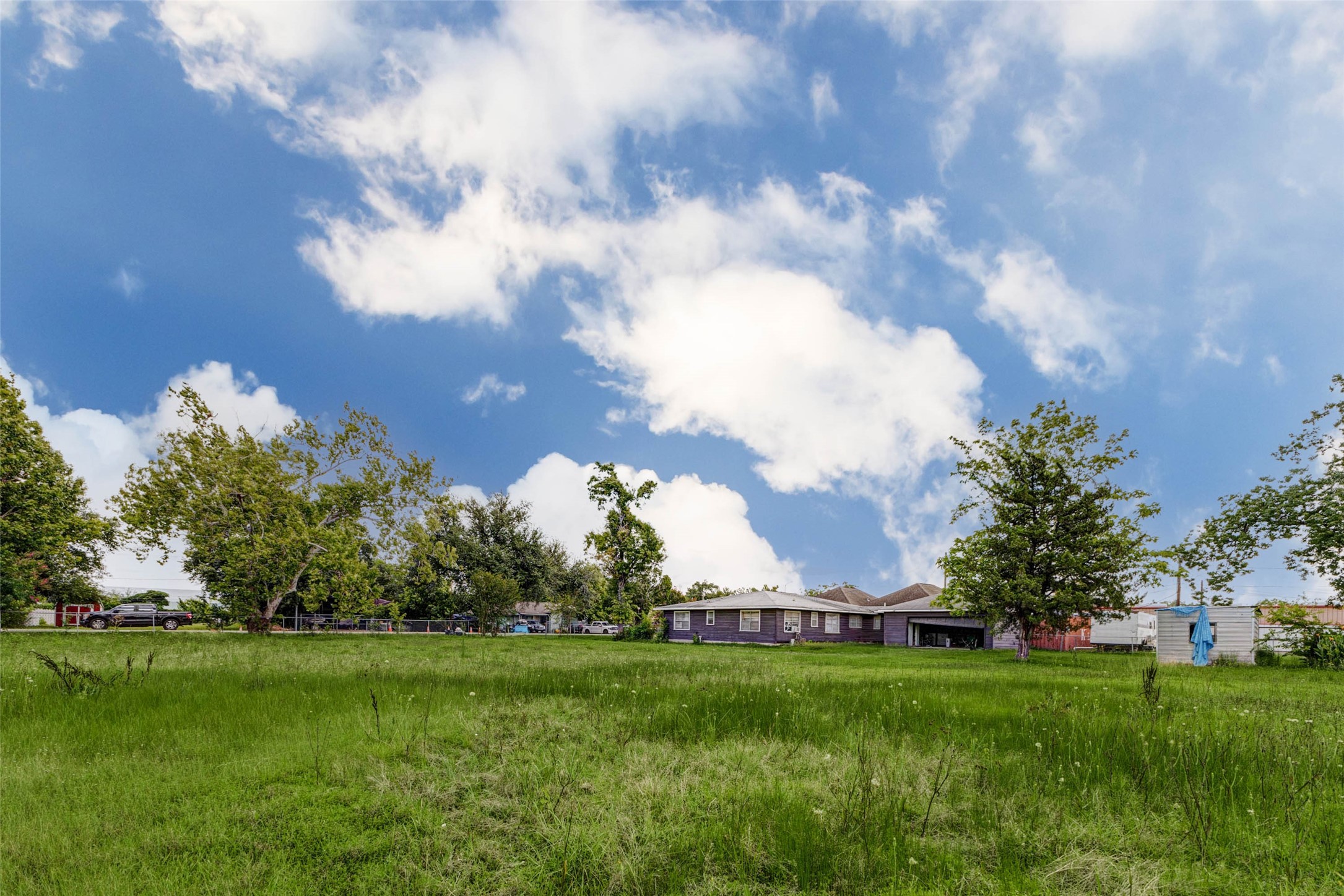 8537 Rannie Road Houston, TX 77080 - Photo 8 of 15 Expansive open-lot view capturing the home and detached garage set back beyond a wide stretch of green space. Mature trees, surrounding fencing, and a bright blue sky frame the property, emphasizing the rare land size and flexible potential of this Spring Branch homesite for outdoor enjoyment, renovation, expansion, investment, or future redevelopment planning.