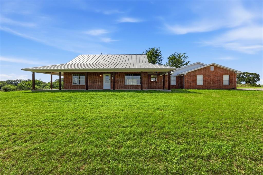 12190 Bell Road Pilot Point, TX 76258 - Photo 3 of 39 Expansive green lawn providing a natural foreground to a brick exterior residence featuring a wide wrap-around porch with supporting pillars, a durable metal roof, and multiple windows