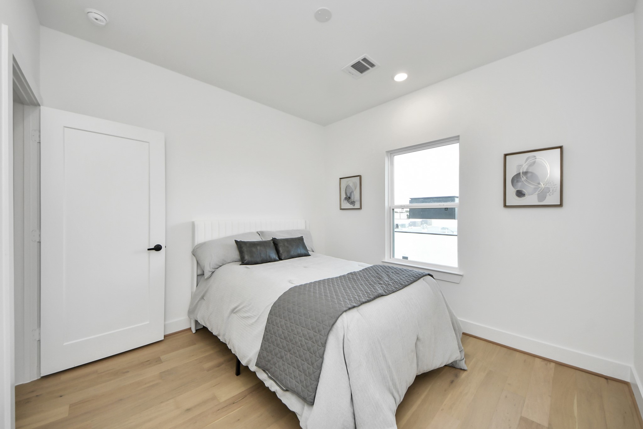 717 Marcella Street, Unit E Houston, TX 77091 - Photo 21 of 25 This is a bright, modern bedroom featuring a comfortable bed with neutral bedding. The room has light wood flooring, white walls, a window providing natural light, and simple wall art, creating a clean and inviting atmosphere.