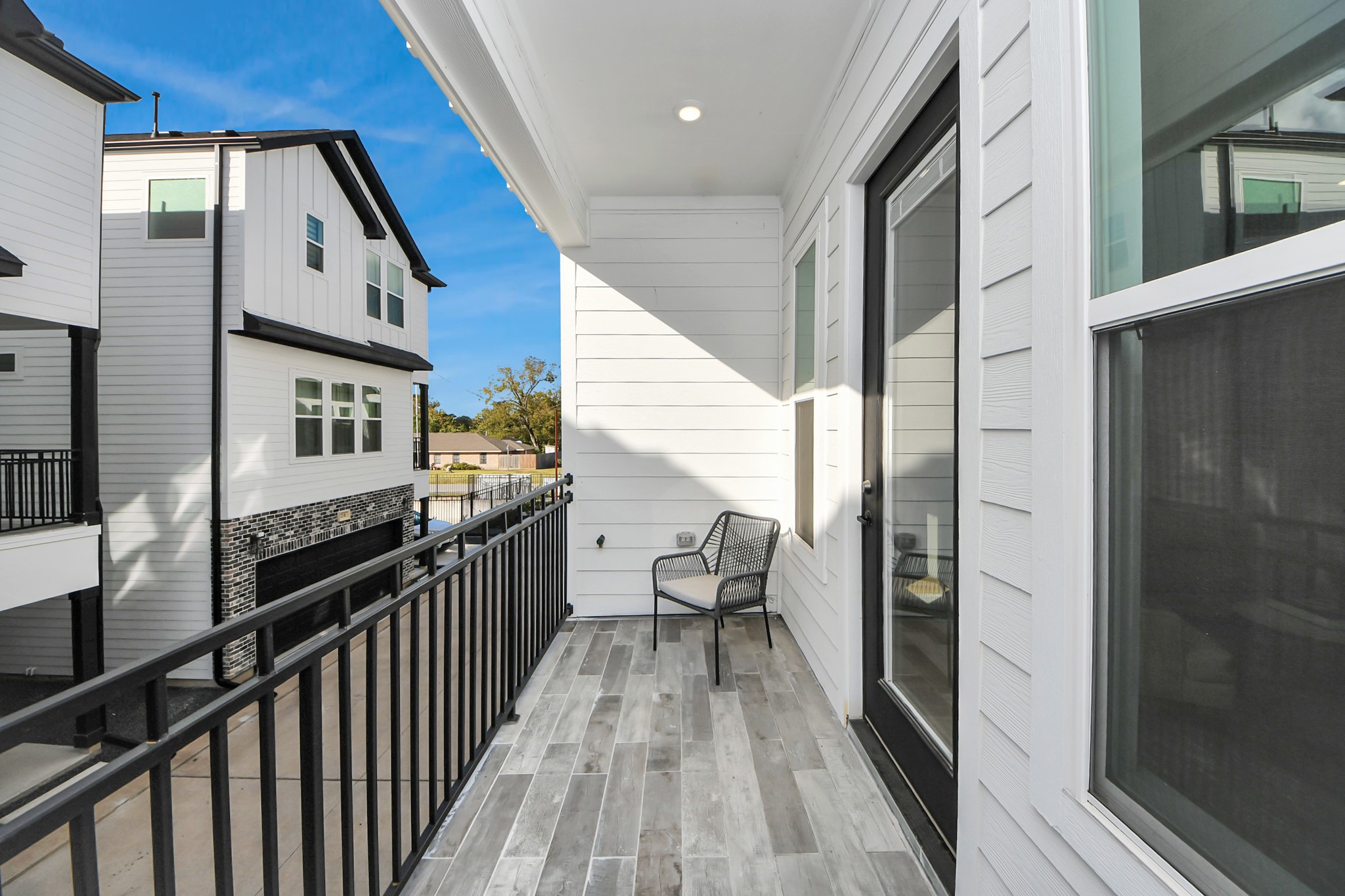 717 Marcella Street, Unit E Houston, TX 77091 - Photo 23 of 25 This photo shows a modern balcony with stylish wood-look tile flooring and a black metal railing. It features a sliding glass door and offers a view of neighboring homes with a similar contemporary design. The space includes a chair, ideal for relaxing outdoors.