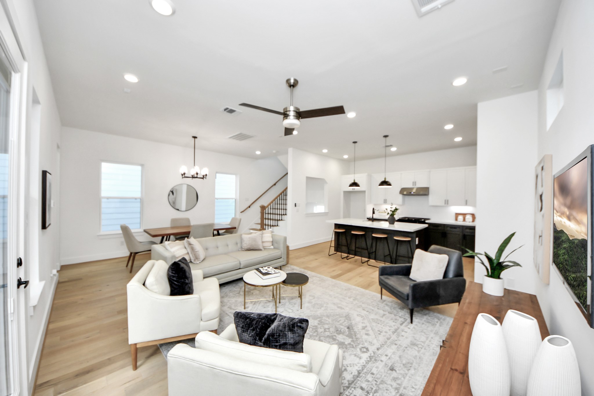 717 Marcella Street, Unit E Houston, TX 77091 - Photo 7 of 25 This photo showcases a modern, open-concept living space featuring a bright living room with a ceiling fan, a dining area, and a sleek kitchen. The kitchen has a large island with barstools, pendant lighting, and white cabinetry. The room is adorned with stylish furniture, wood flooring, and ample natural light.