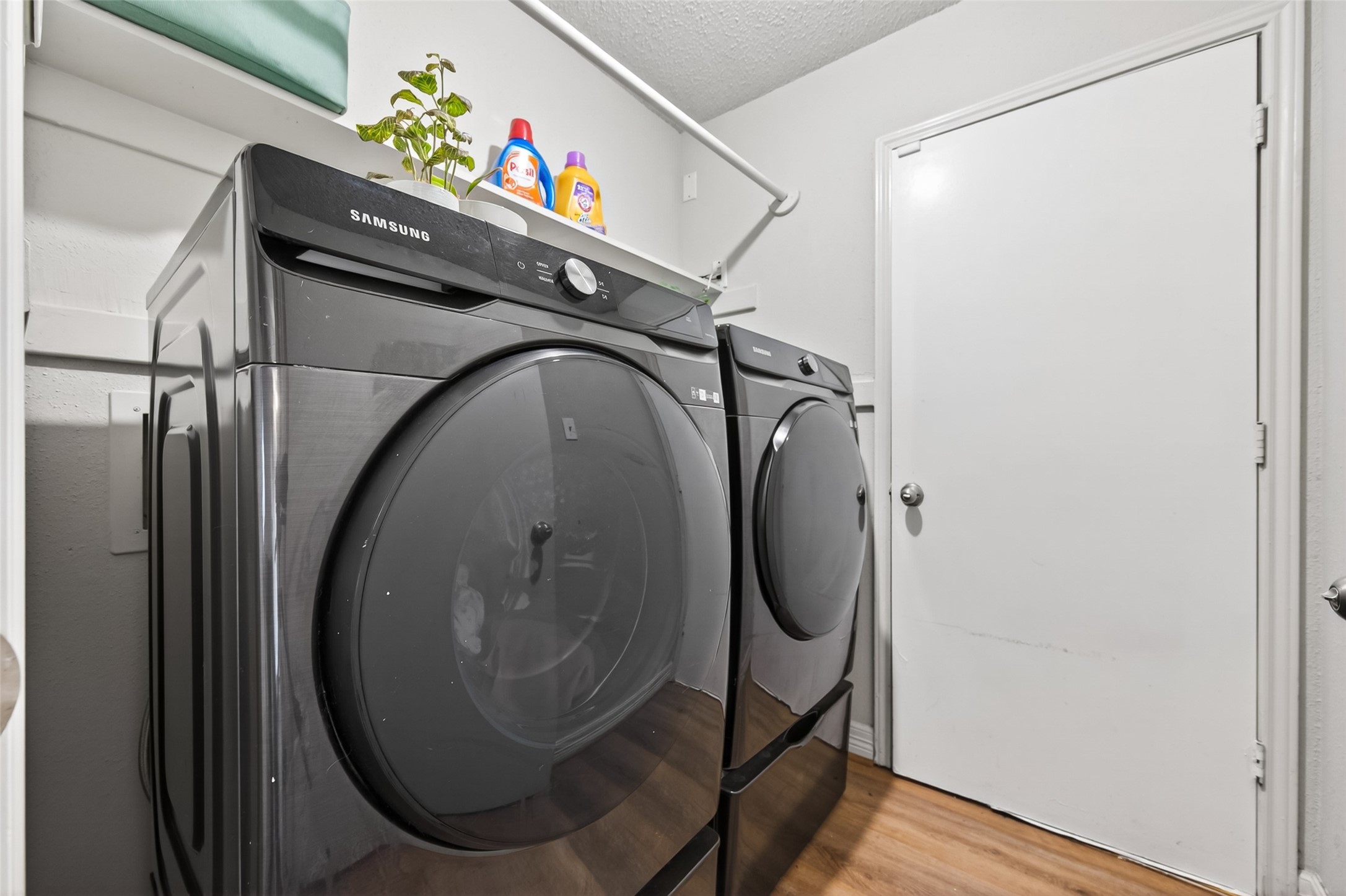 18446 North Willow Bluff Road Katy, TX 77449 - Photo 30 of 33 This photo showcases a laundry room equipped with modern, front-loading Samsung washer and dryer units. The space has a clean, white interior with wooden flooring and a small storage shelf above the appliances.