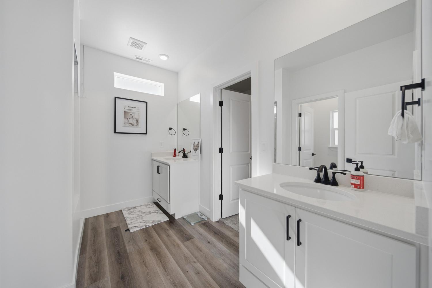 6144 Red Sand Lane St. George, UT 84790 - Photo 14 of 19 Daylight spills through a picture window above this vanity, illuminating the crisp quartz countertop and matte-black faucet finish below. Framed art and a mirrored medicine cabinet add a polished touch, while warm wood-look flooring keeps the space grounded and inviting underfoot.