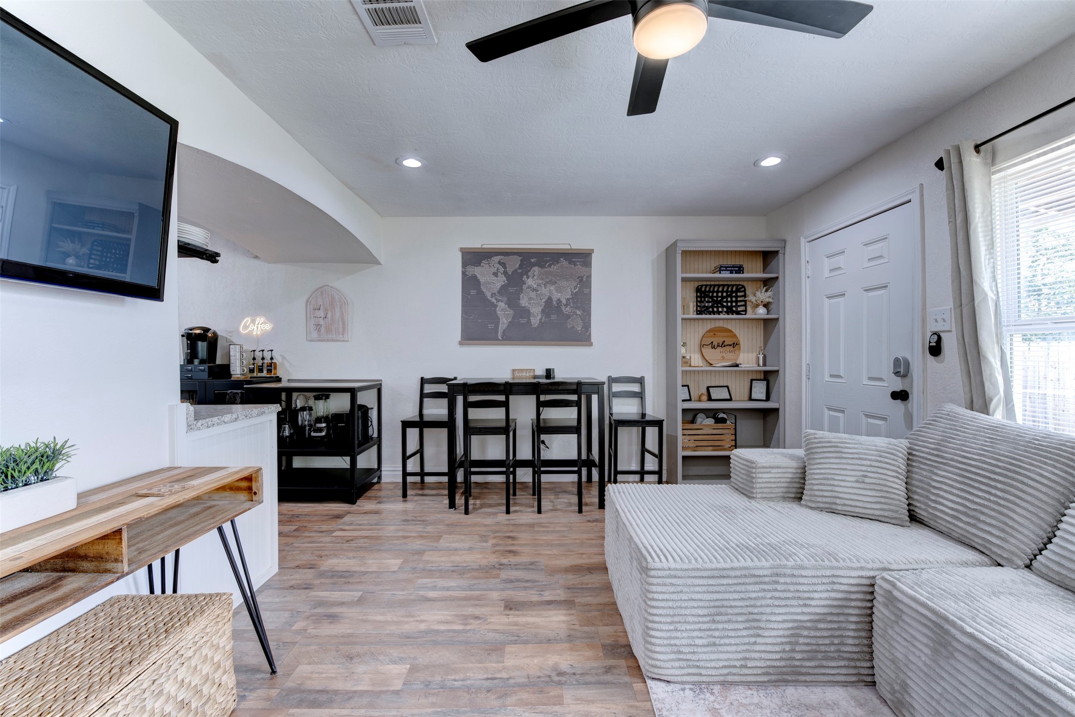 1121 Panama Street Houston, TX 77009 - Photo 8 of 35 Full living and dining layout showing wood-look LVP flooring, five-blade black ceiling fan with light kit, recessed lighting, wall-mounted flat screen TV, built-in five-shelf bookcase with beadboard back panel, black bar-height dining set, dedicated coffee station with neon sign, white six-panel entry door with smart lock, and oversized window with natural light.