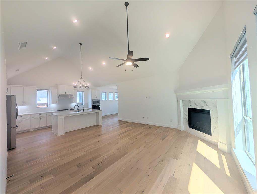 15532 Fringe Tree Road Frisco, TX 75035 - Photo 3 of 31 Open-concept living space featuring vaulted ceilings, light wood-finish flooring, a white kitchen with an island, and a fireplace with a white mantel and marble-look surround