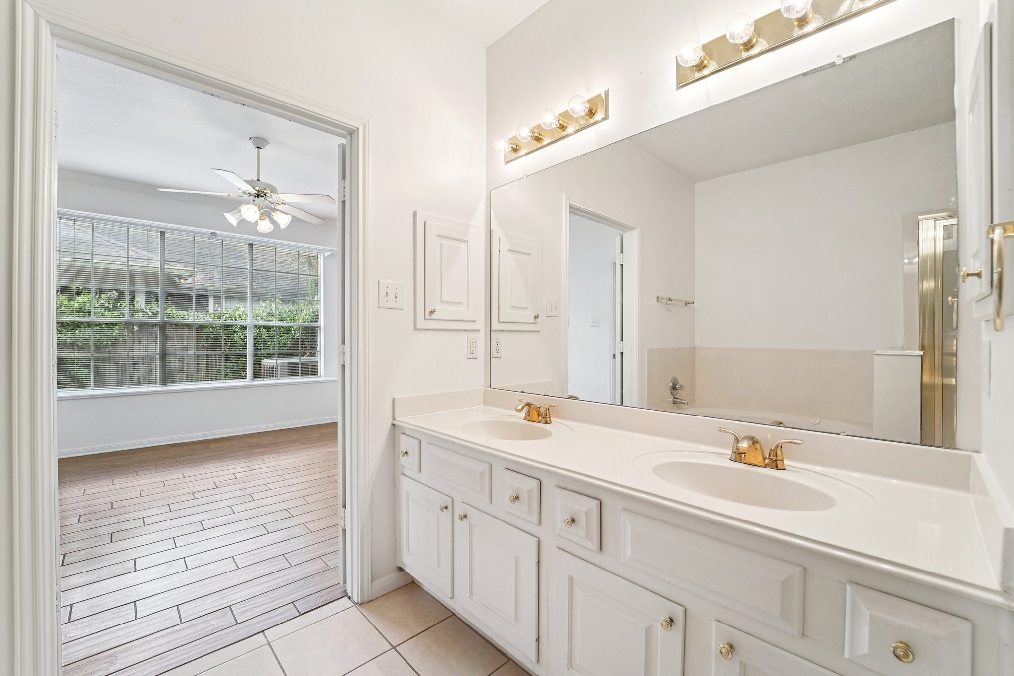 14420 Walters Road, Unit 55 Houston, TX 77014 - Photo 24 of 35 This photo showcases a bright, spacious bathroom with a dual-sink vanity and gold fixtures. It opens to a sunlit room with large windows and wood-style flooring, featuring a ceiling fan for comfort.