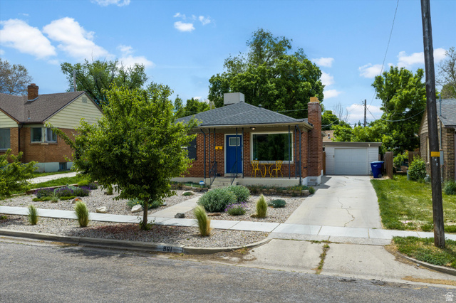 961 33rd Street Ogden, UT 84403 - Photo 33 of 39 Brick facade residence featuring a prominent picture window, a bright blue front door, a concrete driveway, a detached garage, and a low-maintenance xeriscape front yard with mature plantings