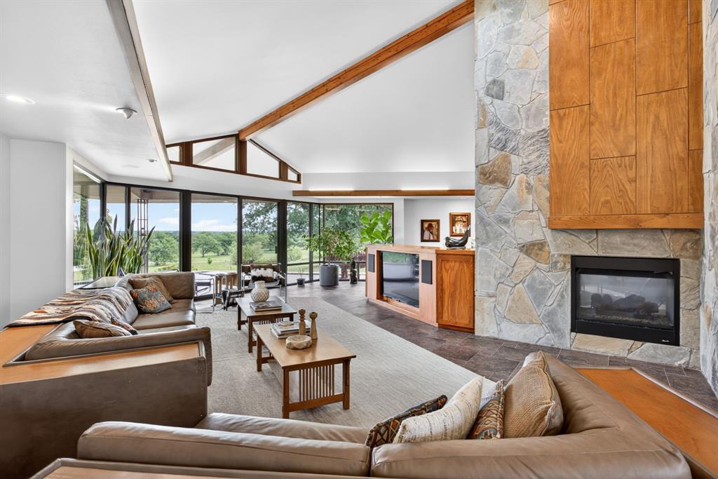 1080 North Greenbrier Road Nocona, TX 76255 - Photo 7 of 40 Expansive living area featuring a stone fireplace with a wood surround, vaulted ceilings with exposed wood beams, and a wall of windows offering landscape views
