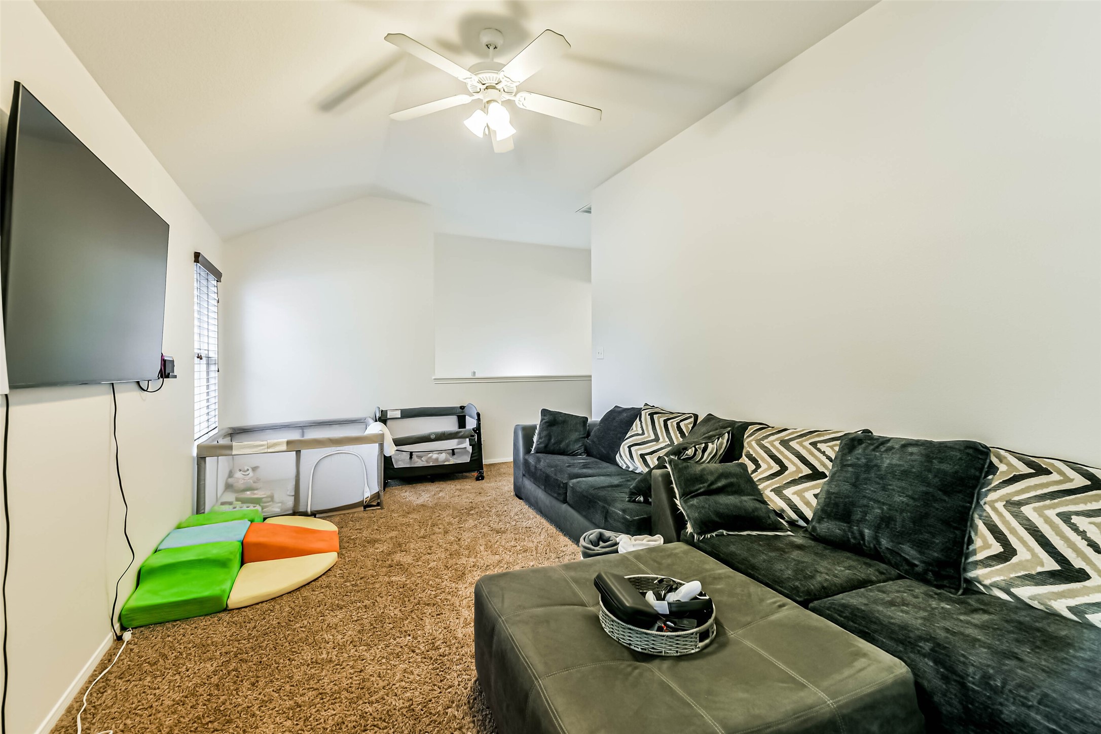 22538 Goss Spring Court Spring, TX 77373 - Photo 22 of 36 This cozy living space features a comfortable sectional sofa with patterned cushions and a ceiling fan for ventilation. A wall-mounted TV offers entertainment, while natural light streams in from a side window. Perfect for relaxation or family gatherings.