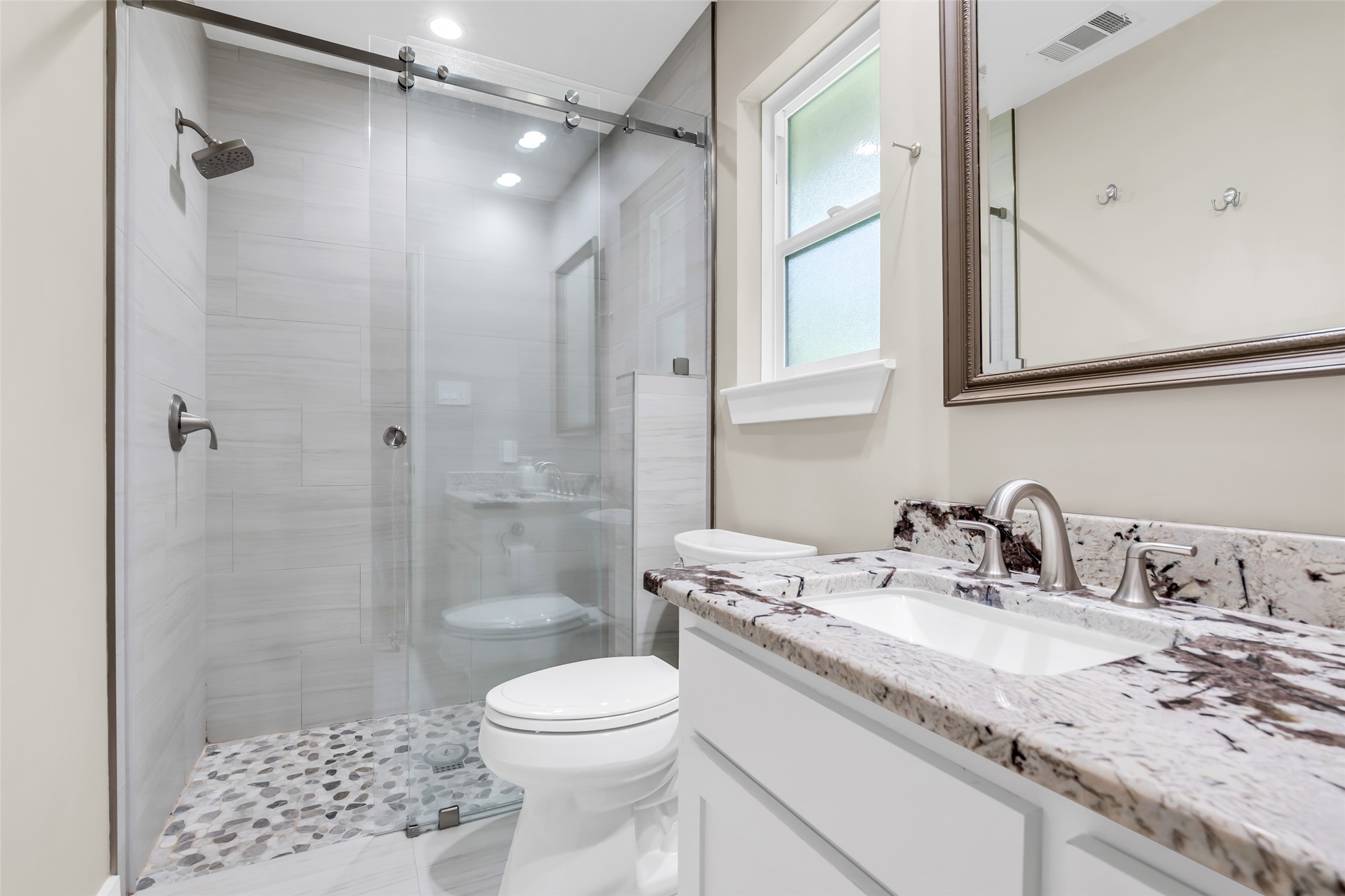22502 Fields Lane Spring, TX 77389 - Photo 20 of 39 This modern bathroom features a sleek glass-enclosed shower with a pebble stone floor, a marble countertop with dual sinks, and a large mirror. The neutral tones and ample lighting create a fresh, inviting space.