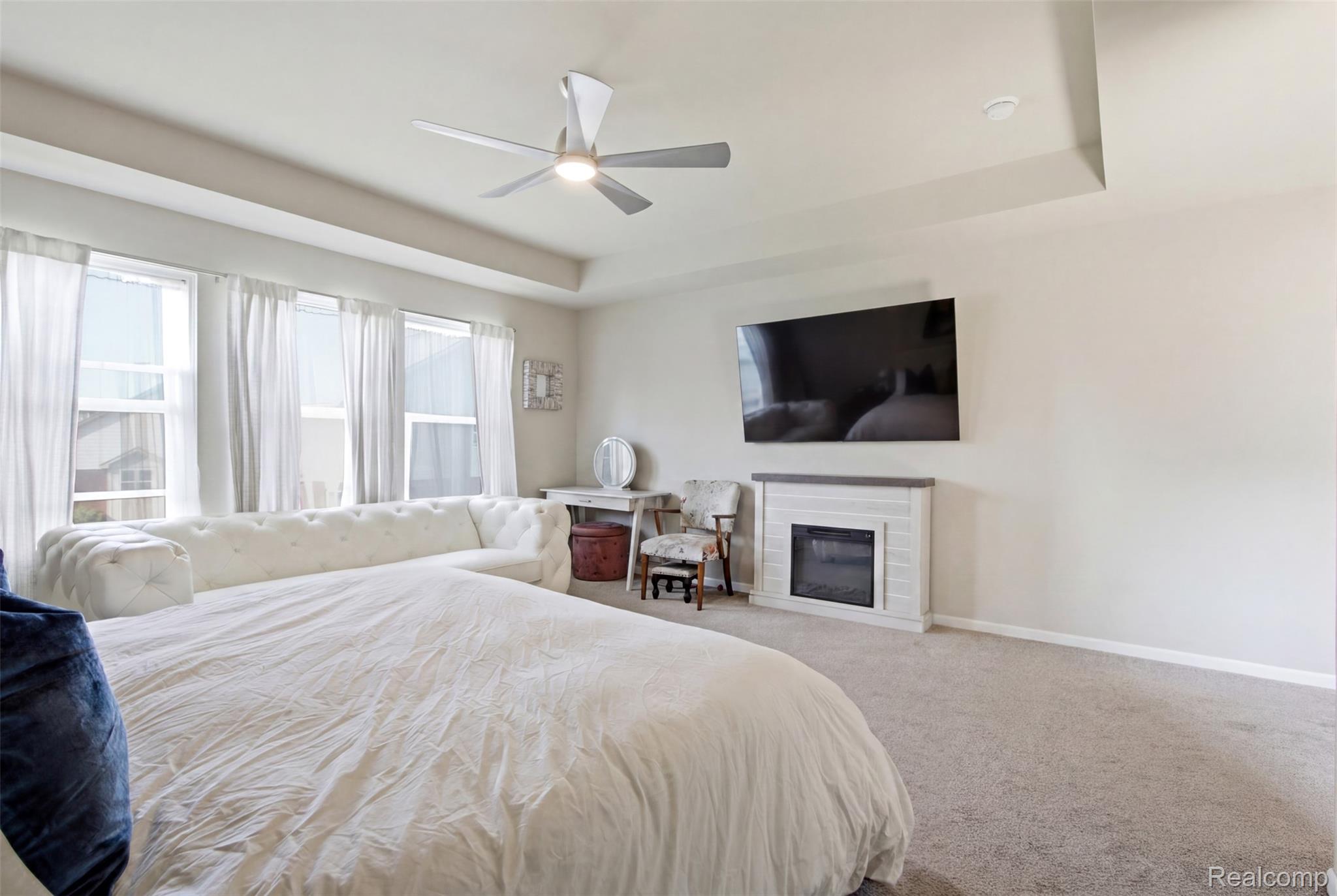 5085 Ridge Lane Warren, MI 48092 - Photo 15 of 29 Carpeted sleeping area featuring a tray ceiling, ceiling fan with integrated lighting, three windows with white drapes, and a built-in fireplace with a tiled surround