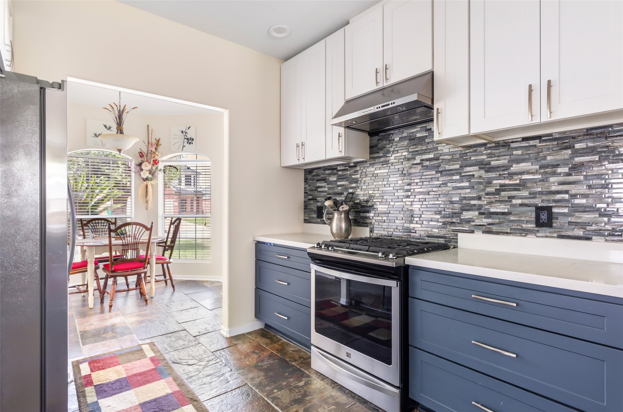 12122 Landsdown Ridge Way Humble, TX 77346 - Photo 14 of 34 Modern kitchen with sleek stainless steel appliances, white and blue 42” cabinetry, and a stylish tile backsplash. A cozy dining area with natural light is visible through an archway, enhancing the inviting atmosphere.