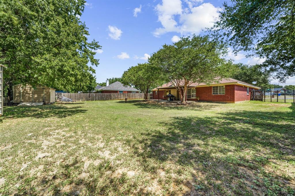 10127 Cougar Ridge Parkway Waco, TX 76708 - Photo 11 of 33 Expansive grass lawn featuring a wood privacy fence, mature trees, a brick-clad home with a covered patio, and a separate wood-sided storage structure