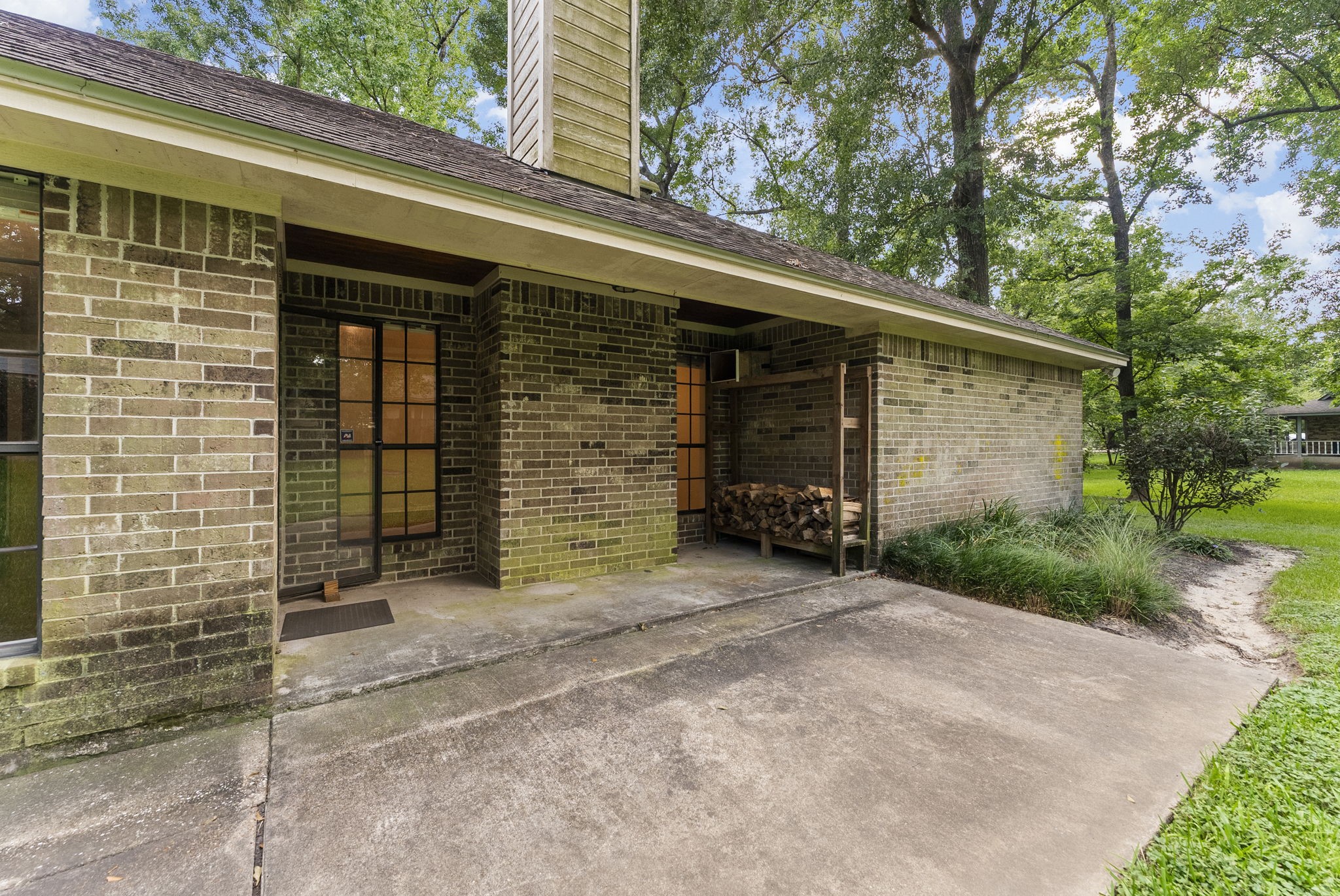 202 Sunset Boulevard Conroe, TX 77303 - Photo 42 of 50 This photo shows a brick exterior of a house with a covered patio area. There's a glass door entrance and a stack of firewood stored neatly. The surrounding area is lush with greenery and tall trees, providing a serene and private setting.