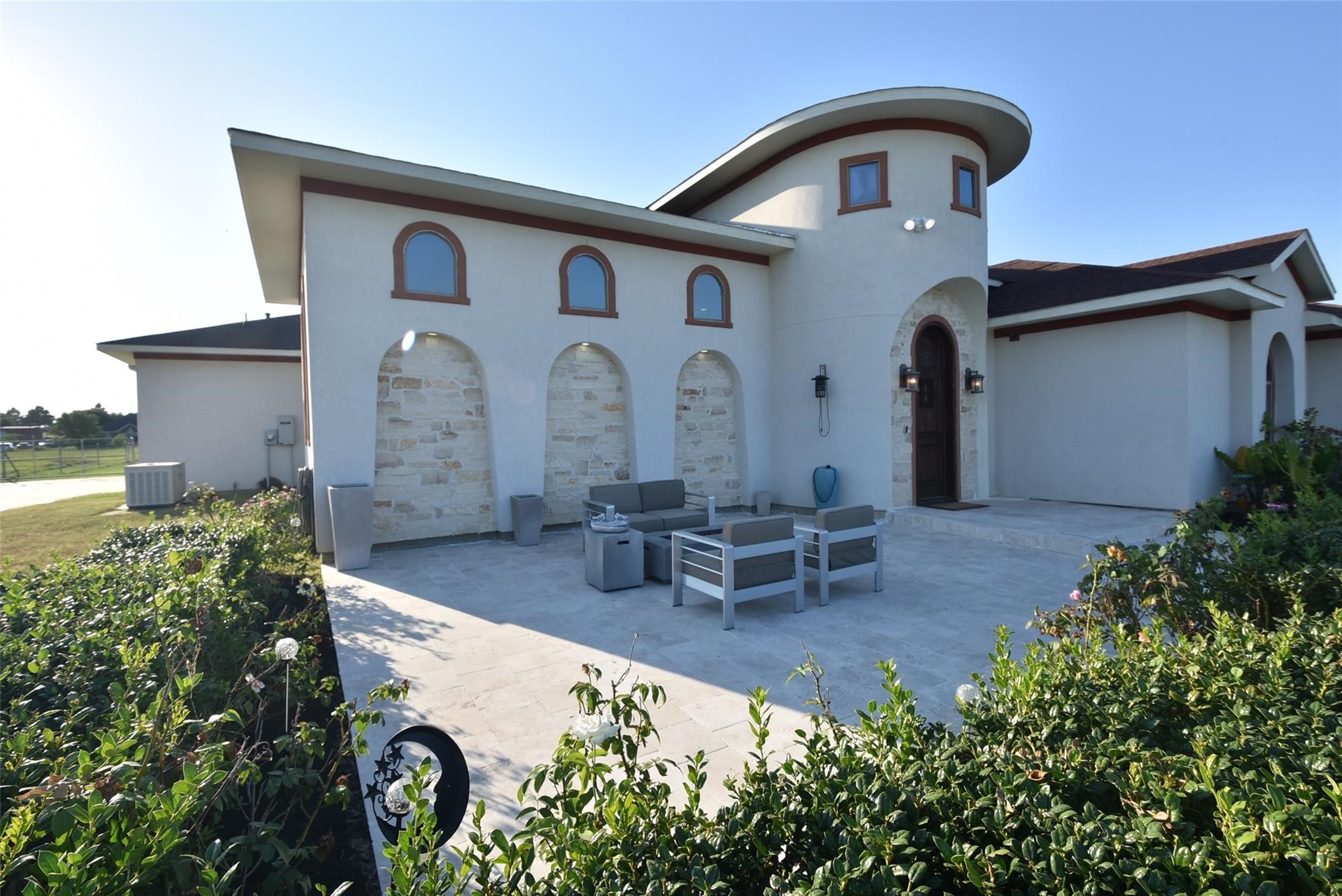 This modern home features a unique architectural design with a rounded tower and arched windows. The exterior is complemented by a spacious stone patio with outdoor seating, surrounded by greenery, offering a serene outdoor space.