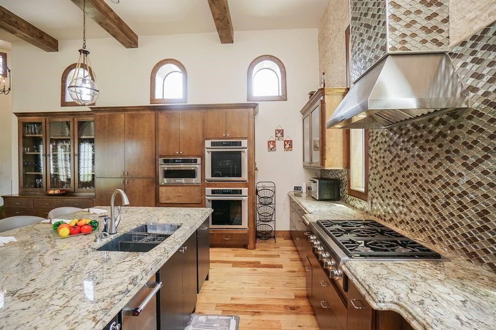 825 Grubbs Road Sealy, TX 77474 - Photo 14 of 47 This modern kitchen features high-end appliances, a spacious granite island with a sink, and elegant wood cabinetry. The room is well-lit with natural light from arched windows and accented with exposed beams and a stylish tiled backsplash.