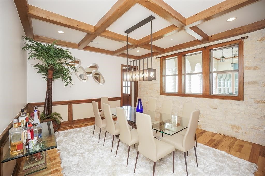 825 Grubbs Road Sealy, TX 77474 - Photo 16 of 47 This dining room features a modern glass table with elegant beige chairs, a textured stone accent wall, and warm wood ceiling beams. A contemporary light fixture and decorative mirrors add style, while a plush rug and a bar area enhance comfort and functionality.