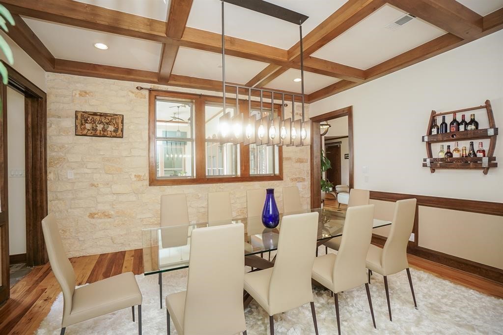 825 Grubbs Road Sealy, TX 77474 - Photo 18 of 47 This dining room features a modern glass table with beige chairs, a coffered ceiling with wood beams, stone accent wall, and contemporary pendant lighting. A small bar shelf and warm wood flooring add elegance to the space.