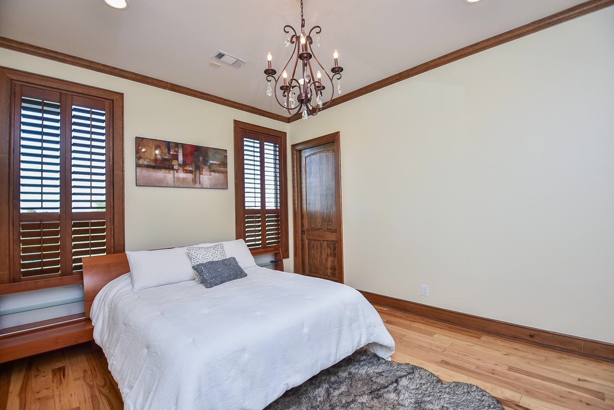825 Grubbs Road Sealy, TX 77474 - Photo 25 of 47 This bedroom features hardwood flooring, a rustic wooden bed frame, and elegant wood trim. It has plantation shutters, a cozy rug, and a decorative chandelier, creating a warm and inviting atmosphere.