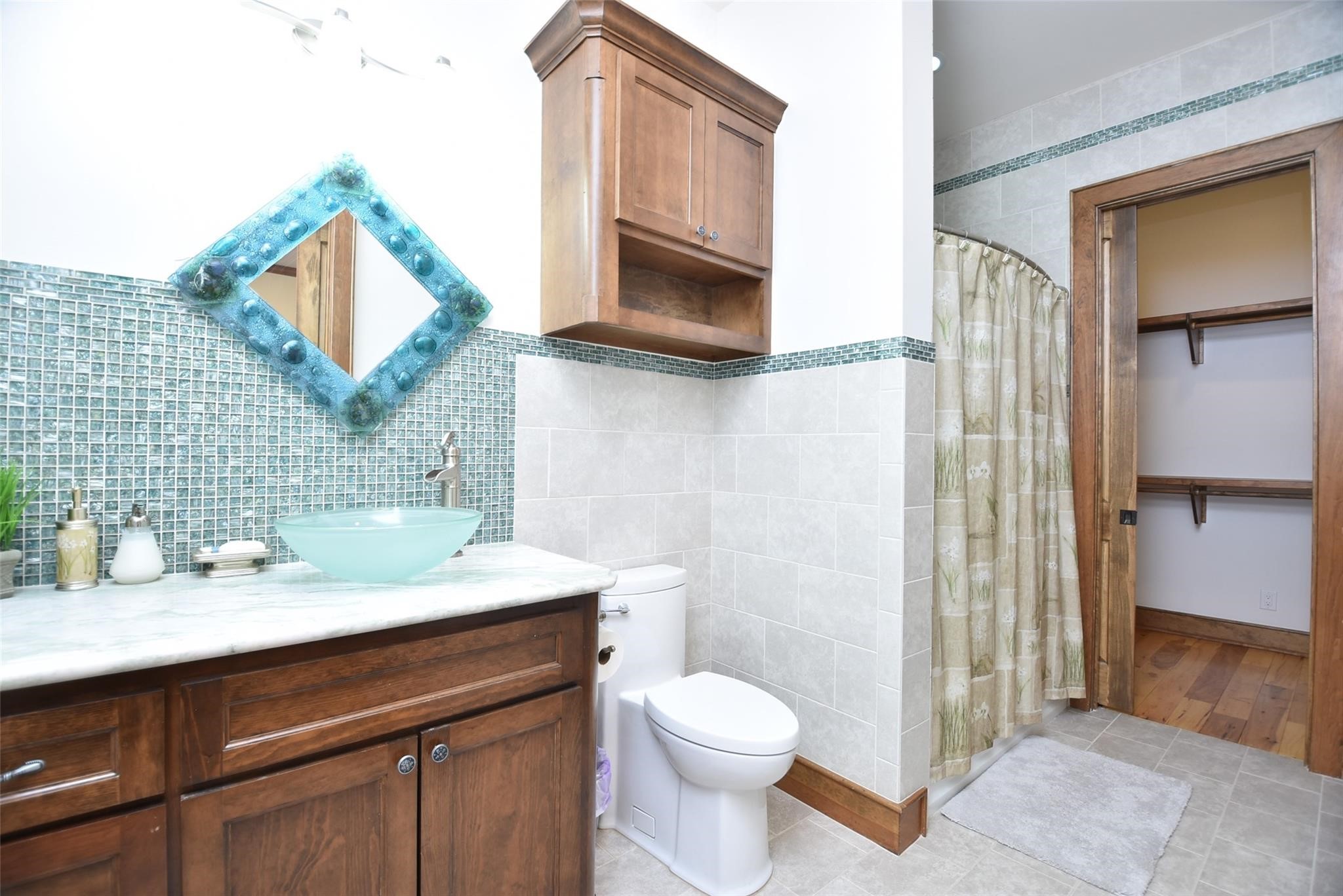 825 Grubbs Road Sealy, TX 77474 - Photo 26 of 47 This bathroom features elegant wood cabinetry, a stylish glass vessel sink, and a unique blue-tiled mirror. It has a tiled shower area with a curtain and an adjacent walk-in closet, providing ample storage space.
