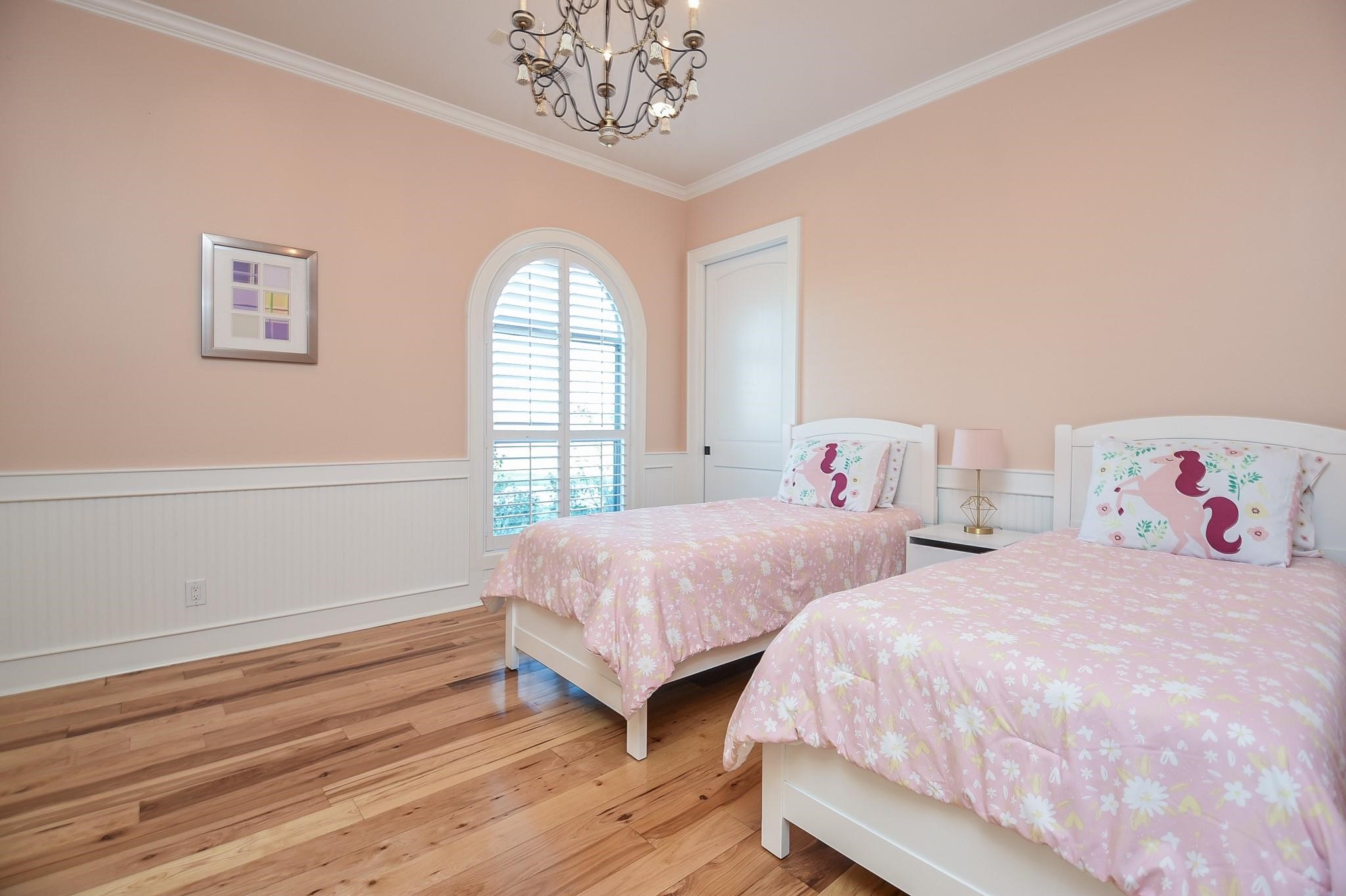 825 Grubbs Road Sealy, TX 77474 - Photo 27 of 47 This cozy bedroom features twin beds with playful pink bedding, a stylish chandelier, and an arched window with shutters. The room is accented with soft peach walls and wood flooring, creating a warm, inviting space.