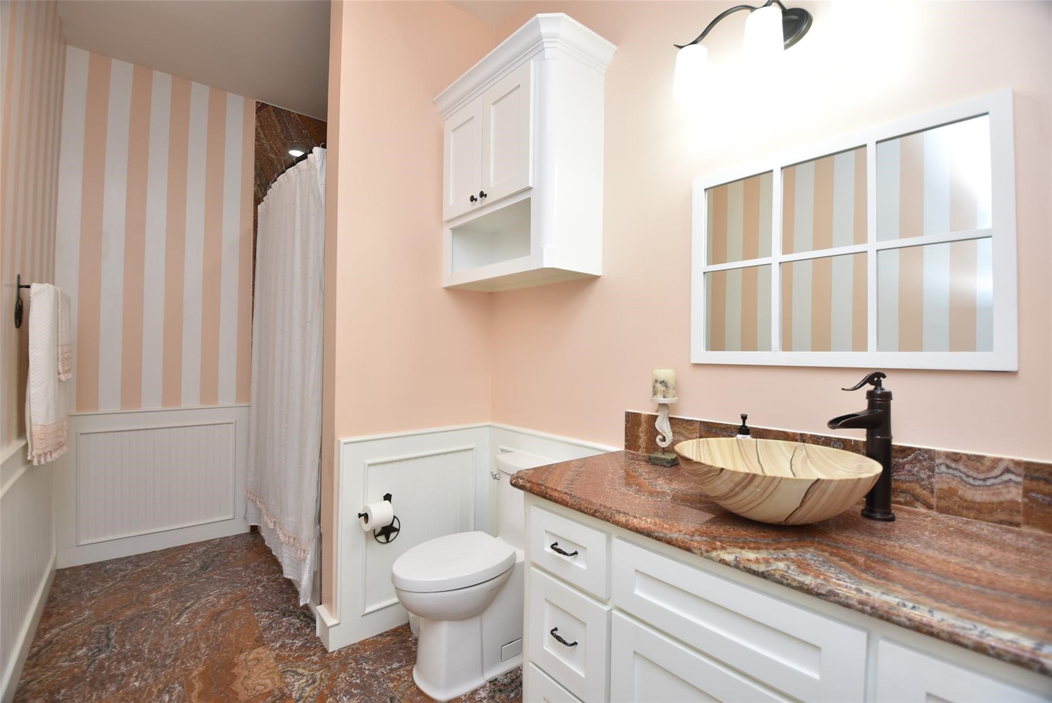 825 Grubbs Road Sealy, TX 77474 - Photo 28 of 47 This bathroom features a stylish design with warm peach tones and white accents. It includes a vessel sink with a unique faucet, spacious granite countertop, striped wallpaper, and a shower with a curtain. The cabinetry offers ample storage, and the lighting is bright and welcoming.
