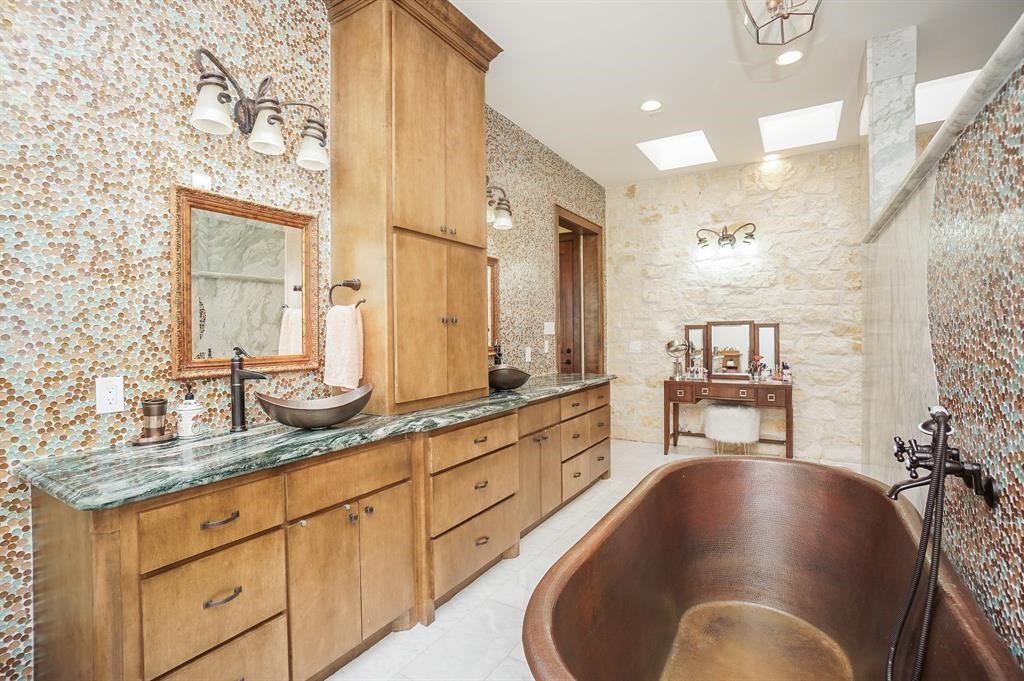 825 Grubbs Road Sealy, TX 77474 - Photo 32 of 47 This luxurious bathroom features a striking copper bathtub and a long vanity with dual vessel sinks set on a green marble countertop. The room is adorned with mosaic tile walls, wood cabinetry, and ample natural light from skylights, creating an elegant and inviting atmosphere.