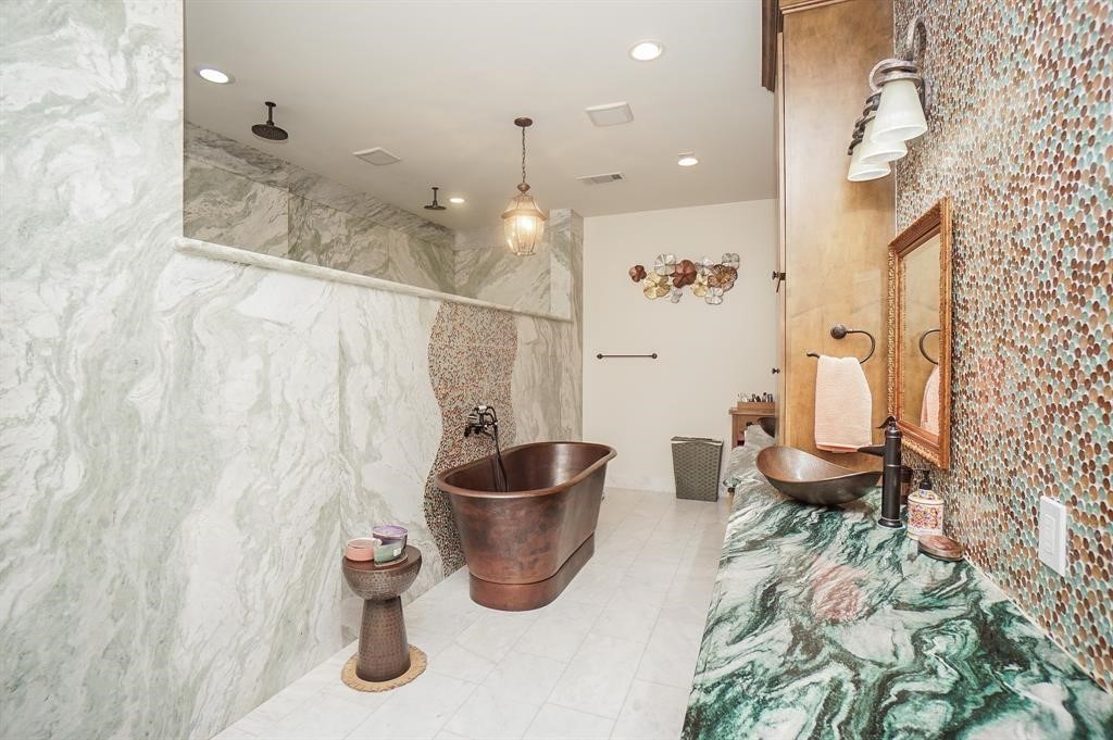 825 Grubbs Road Sealy, TX 77474 - Photo 35 of 47 This bathroom features a luxurious copper bathtub set against elegant marble walls. It includes a spacious, walk-in shower with dual showerheads. The countertop is a striking green marble with a unique vessel sink. Warm lighting and textured wall accents enhance the sophisticated ambiance.