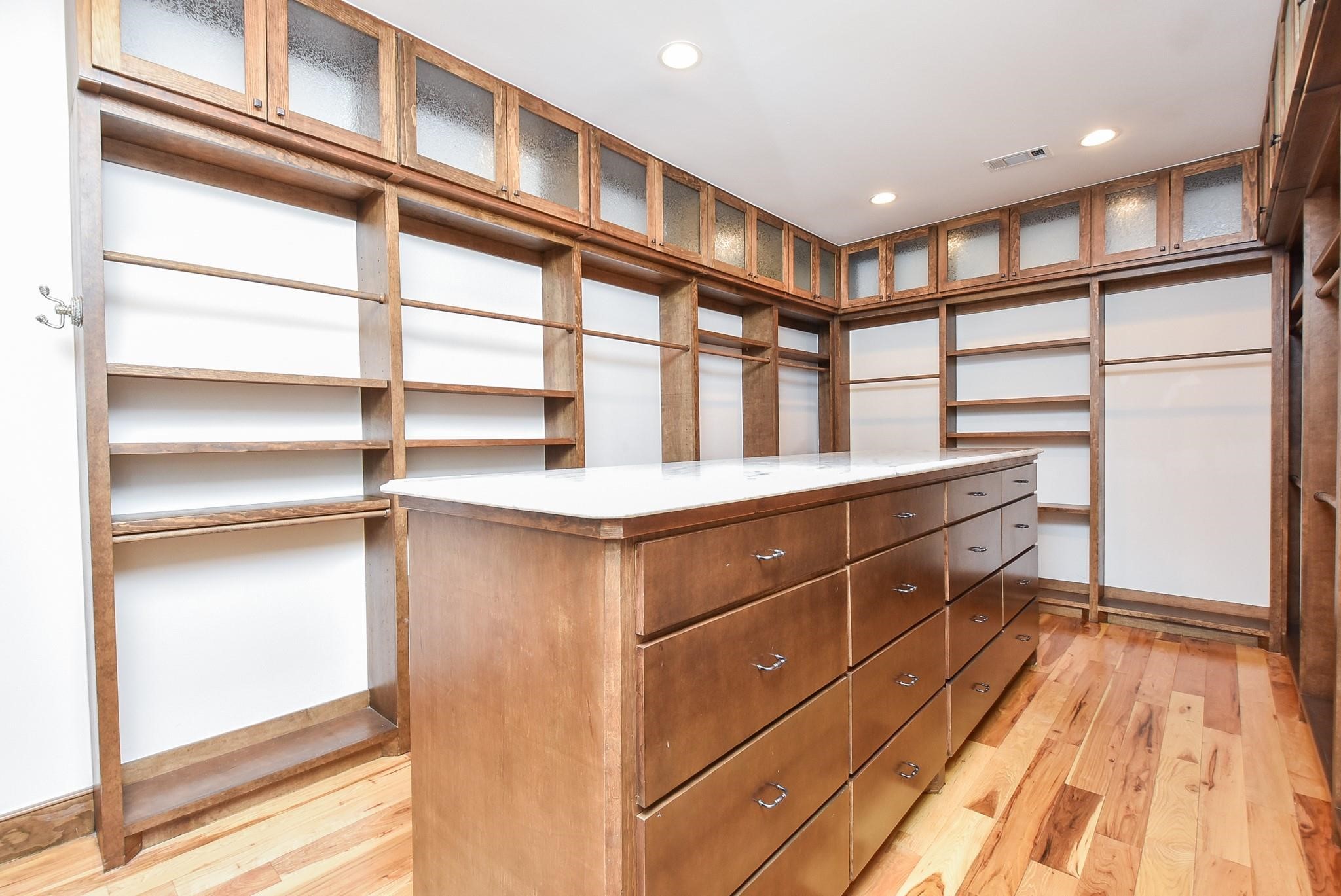 825 Grubbs Road Sealy, TX 77474 - Photo 36 of 47 This spacious walk-in closet features custom wood shelving, ample hanging space, and a large central island with numerous drawers, perfect for organization. The hardwood floors and overhead lighting add a touch of elegance.