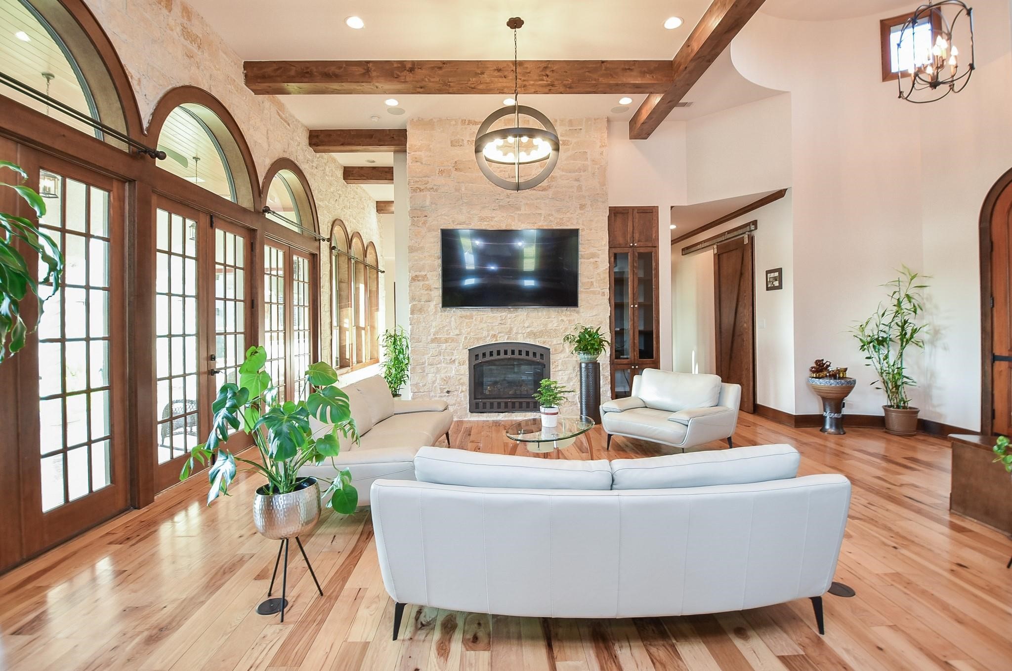 825 Grubbs Road Sealy, TX 77474 - Photo 8 of 47 This living room features high ceilings with exposed wooden beams, a stone fireplace, and a wall-mounted TV. Large arched windows provide ample natural light, complemented by elegant French doors. The space is adorned with modern furniture and lush indoor plants, creating a bright and inviting atmosphere.