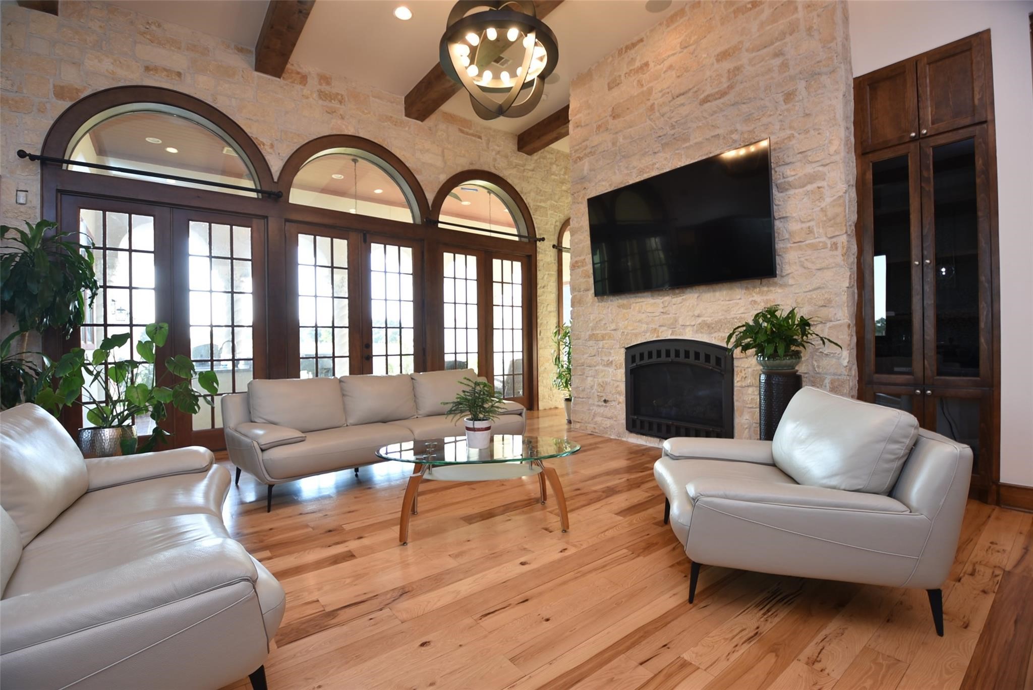 825 Grubbs Road Sealy, TX 77474 - Photo 9 of 47 This inviting living room features elegant wood flooring, high ceilings with exposed beams, and a stone accent wall with a fireplace. Large arched windows provide ample natural light, while modern leather seating and a sleek glass coffee table complete the sophisticated look.