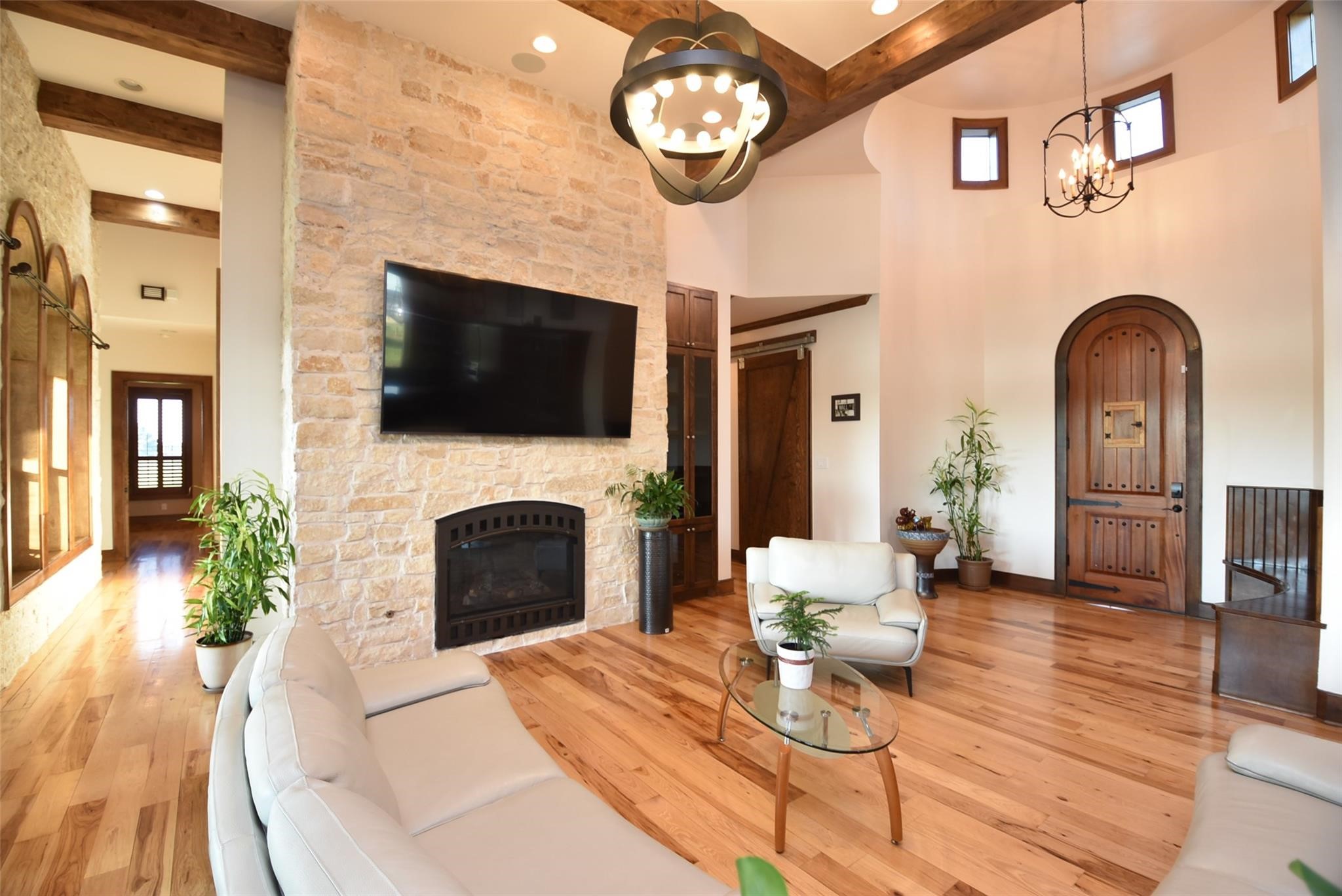 825 Grubbs Road Sealy, TX 77474 - Photo 10 of 47 This inviting living room features a cozy stone fireplace, high ceilings with wooden beams, and a large wall-mounted TV. The space is brightened by natural light and includes elegant furnishings, plants, and a distinctive wooden entrance door, creating a warm and welcoming atmosphere.