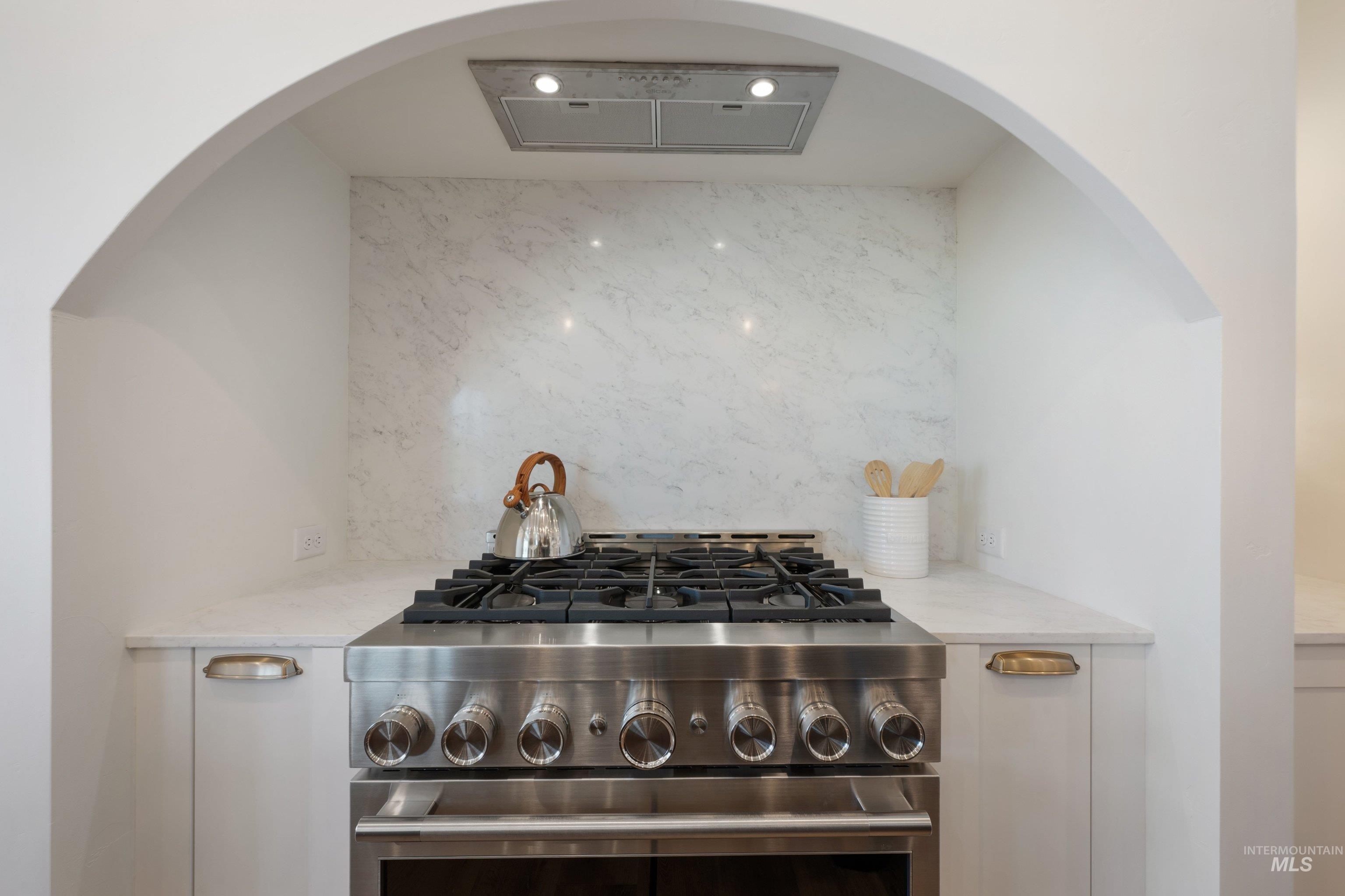 24821 Ardmore Court Caldwell, ID 83607 - Photo 20 of 43 Stainless steel range with a built-in oven, marble-patterned backsplash, white cabinetry with brass hardware, and an integrated range hood