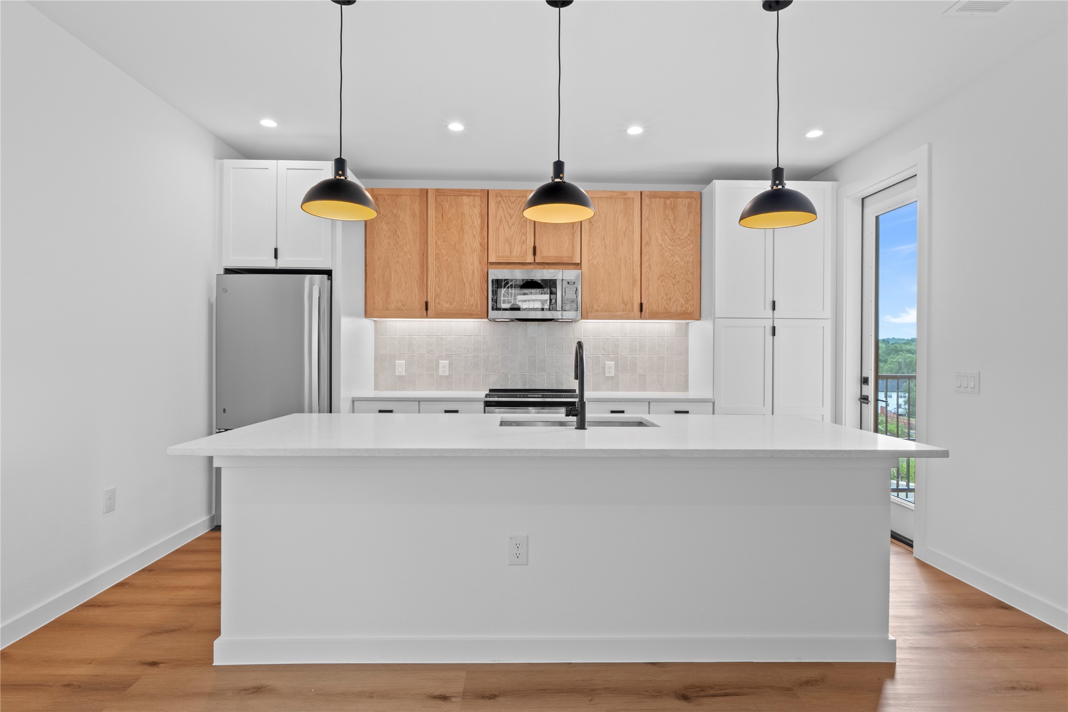 1406 Mason Avenue Austin, TX 78721 - Photo 2 of 36 MODEL HOME - REPRESENTATIVE PHOTO: Modern kitchen featuring a large central island with a solid surface countertop, stainless steel appliances, two-tone cabinetry, wood-finish flooring, and a glass-paneled door leading to an exterior patio.