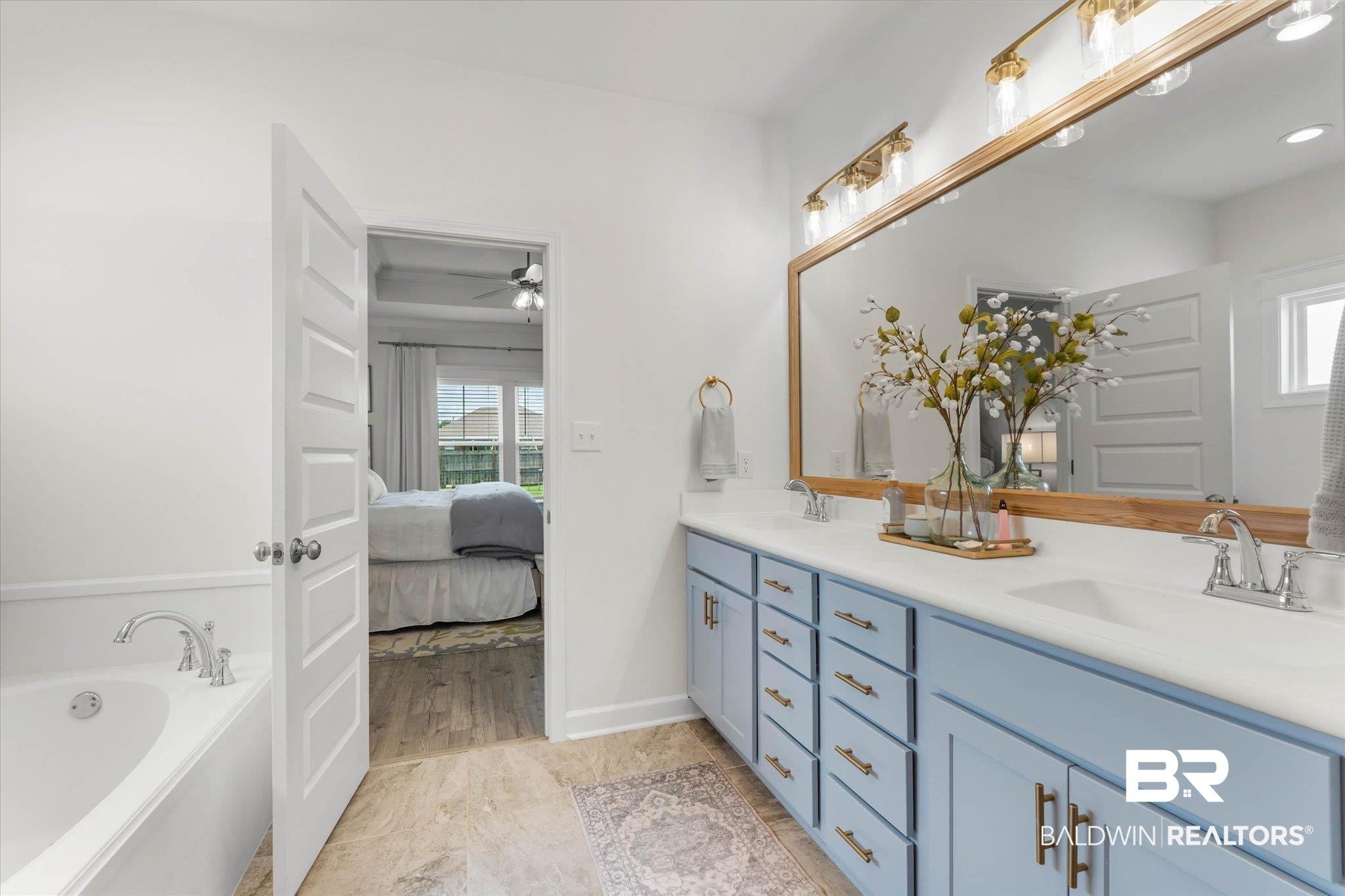 9074 Moses Way Theodore, AL 36582 - Photo 25 of 52 Bathroom featuring a double vanity with light blue cabinetry and brushed gold hardware, a spacious soaking tub, and a large wood-framed mirror with overhead lighting