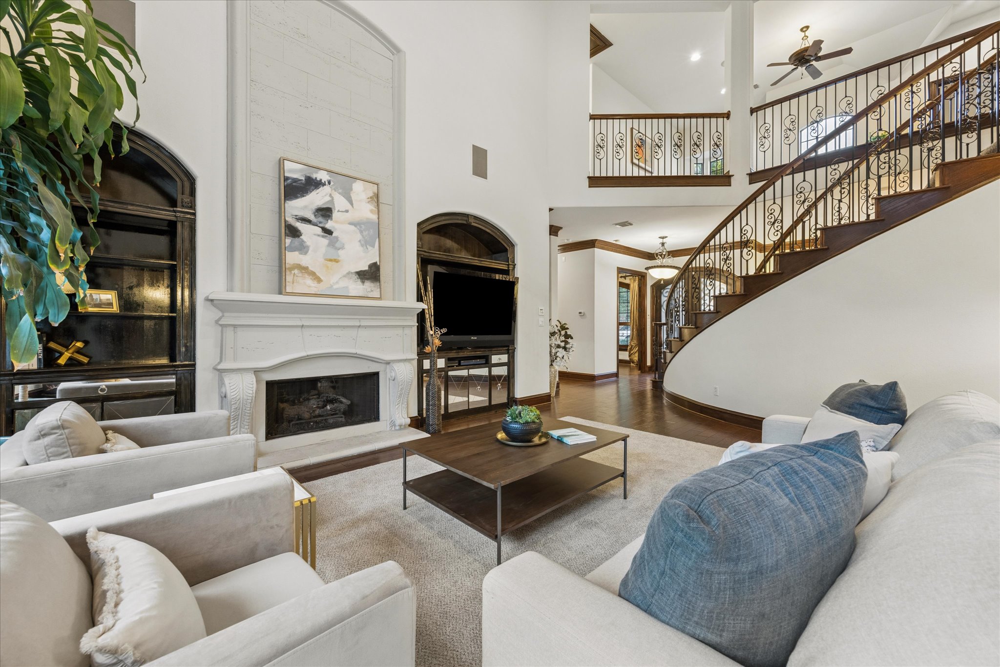Two-story living area featuring a prominent fireplace with a substantial mantel, dark wood-finish flooring, a grand curved staircase with ornate iron balusters, and a built-in arched display cabinet