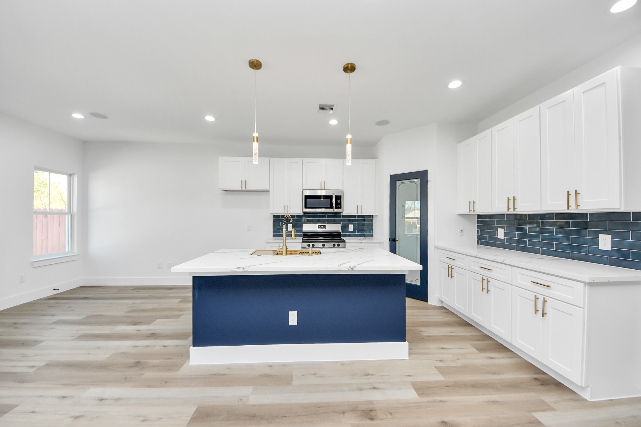 4606 Mallow Street Houston, TX 77051 - Photo 14 of 31 This modern kitchen features a spacious open layout with light flooring. It has a large island with a navy blue base and white countertops. The kitchen boasts white cabinetry with gold handles and a stylish blue tile backsplash. Stainless steel appliances and pendant lighting add to the contemporary design.