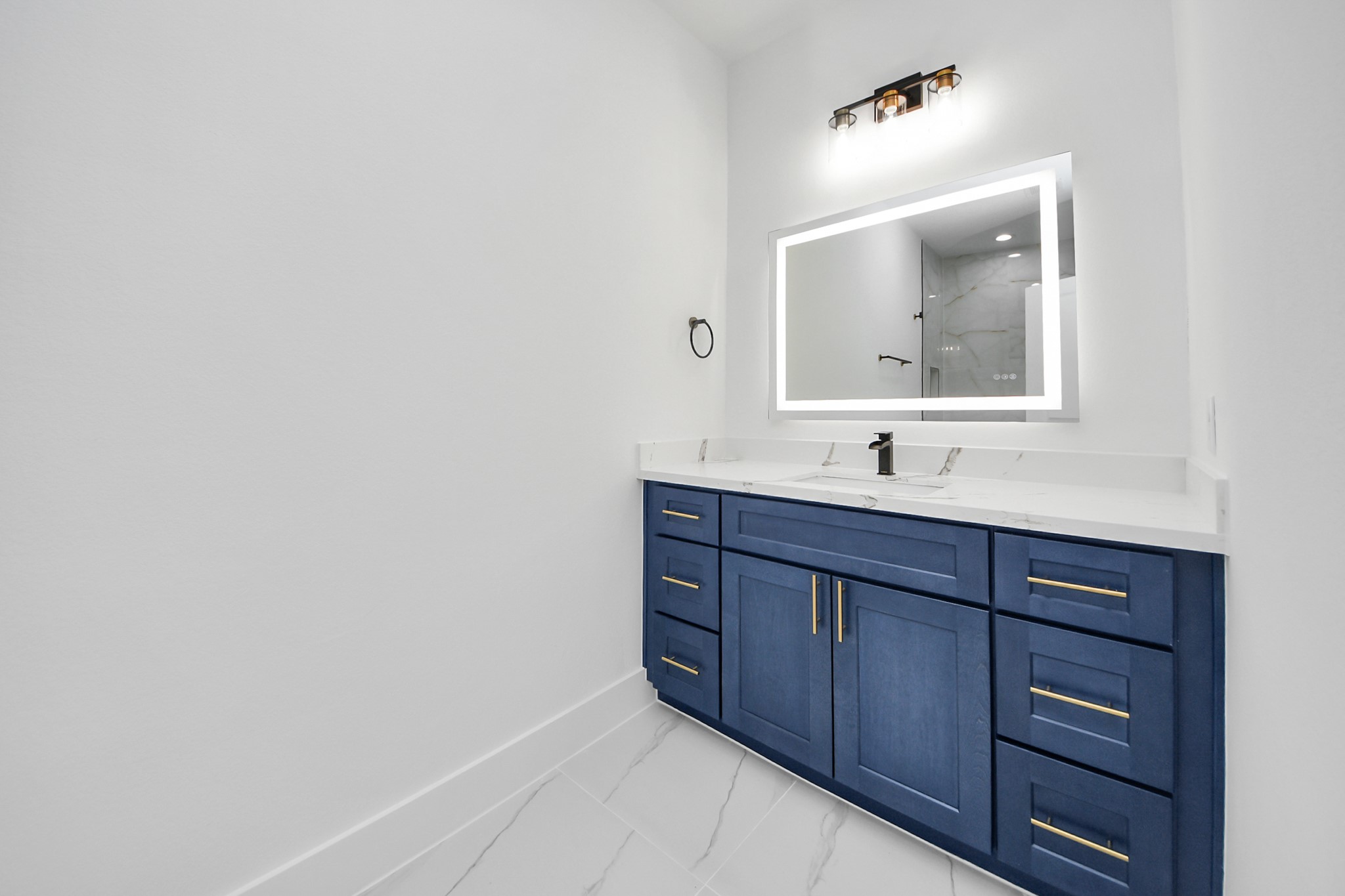 4606 Mallow Street Houston, TX 77051 - Photo 24 of 31 This primary bathroom features a modern design with a striking blue vanity, gold hardware, and a sleek white countertop. The illuminated mirror adds elegance, while the neutral walls and flooring offer a clean, spacious feel.
