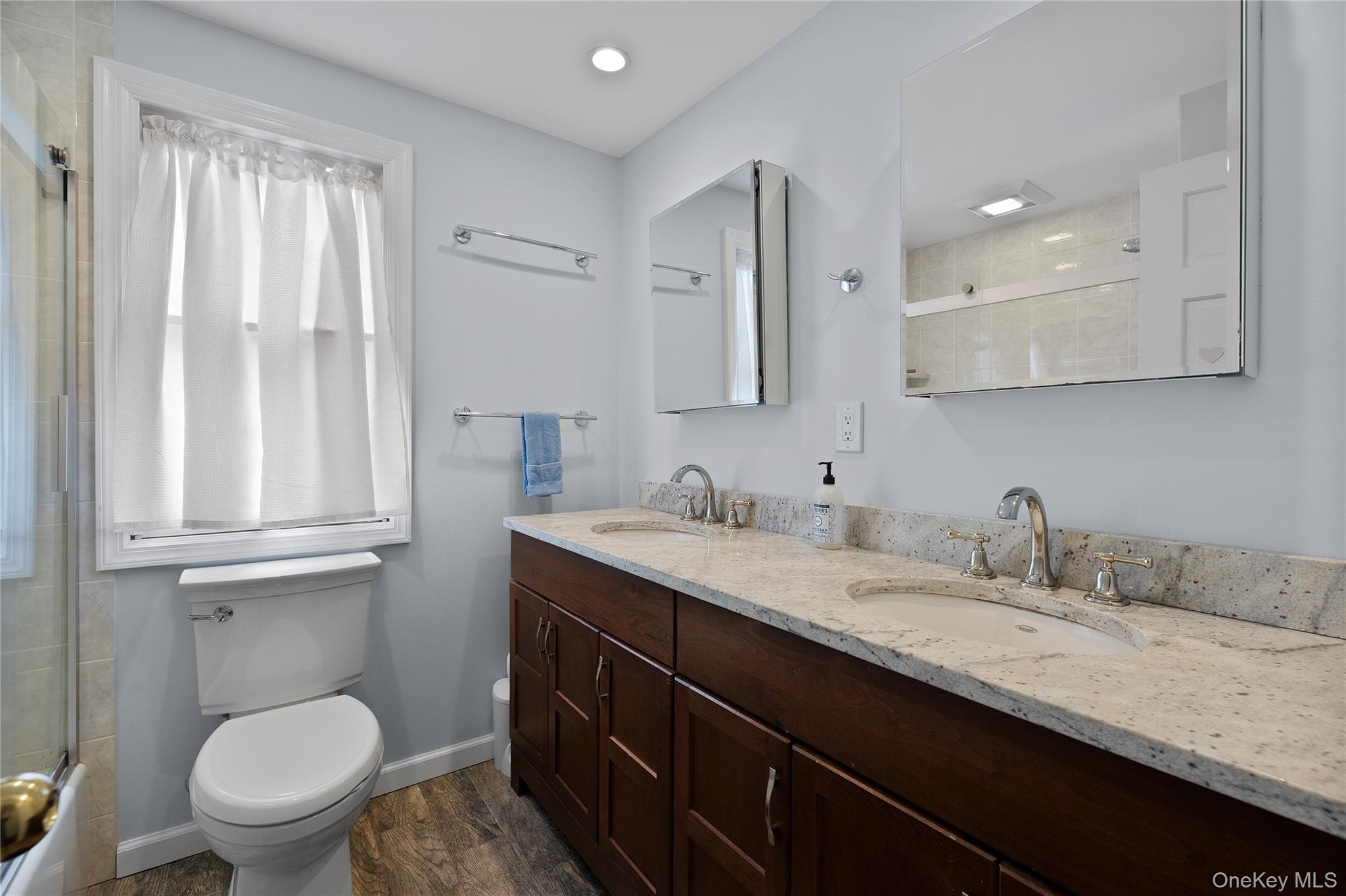 9 Three Sisters Road St. James, NY 11780 - Photo 18 of 36 Guest bathroom featuring a dual vanity with light speckled countertops and undermount sinks, wood-finish cabinetry, two mirrored medicine cabinets, and wood-finish flooring
