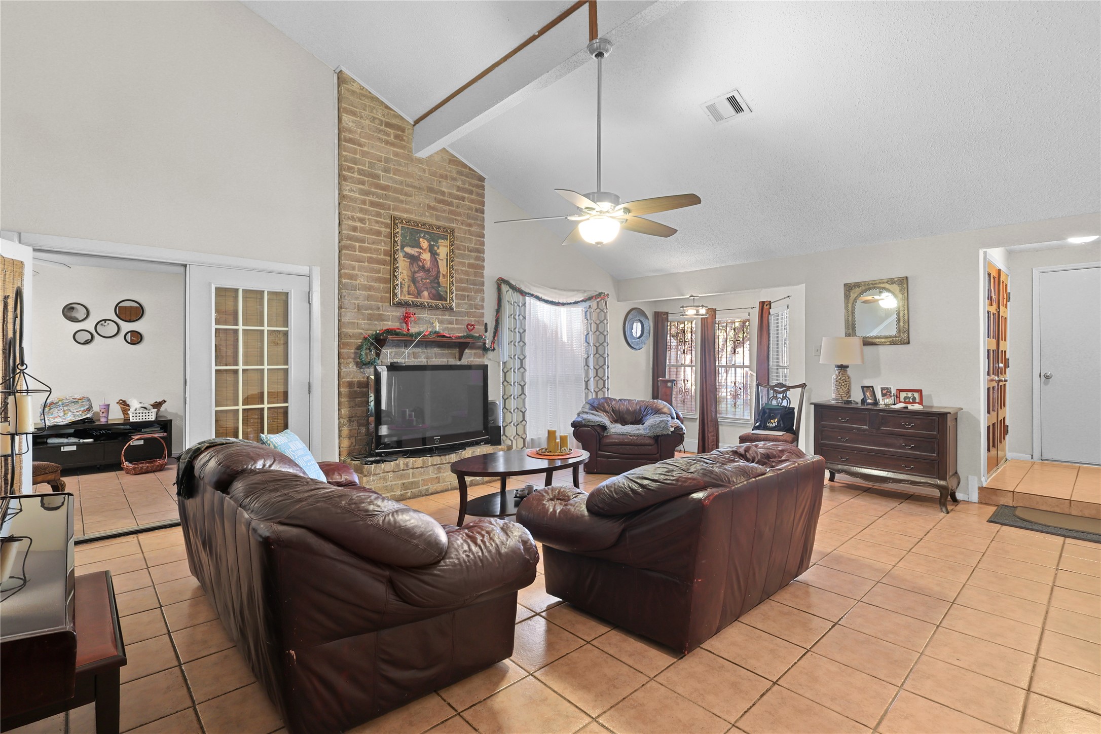 4123 Yupon Ridge Drive Houston, TX 77072 - Photo 20 of 22 This spacious living room features a high vaulted ceiling with a ceiling fan, a cozy brick fireplace, and ample natural light from large windows. It offers a comfortable seating area with leather couches, tile flooring, and access to an adjacent room through French doors, creating a welcoming and open atmosphere.